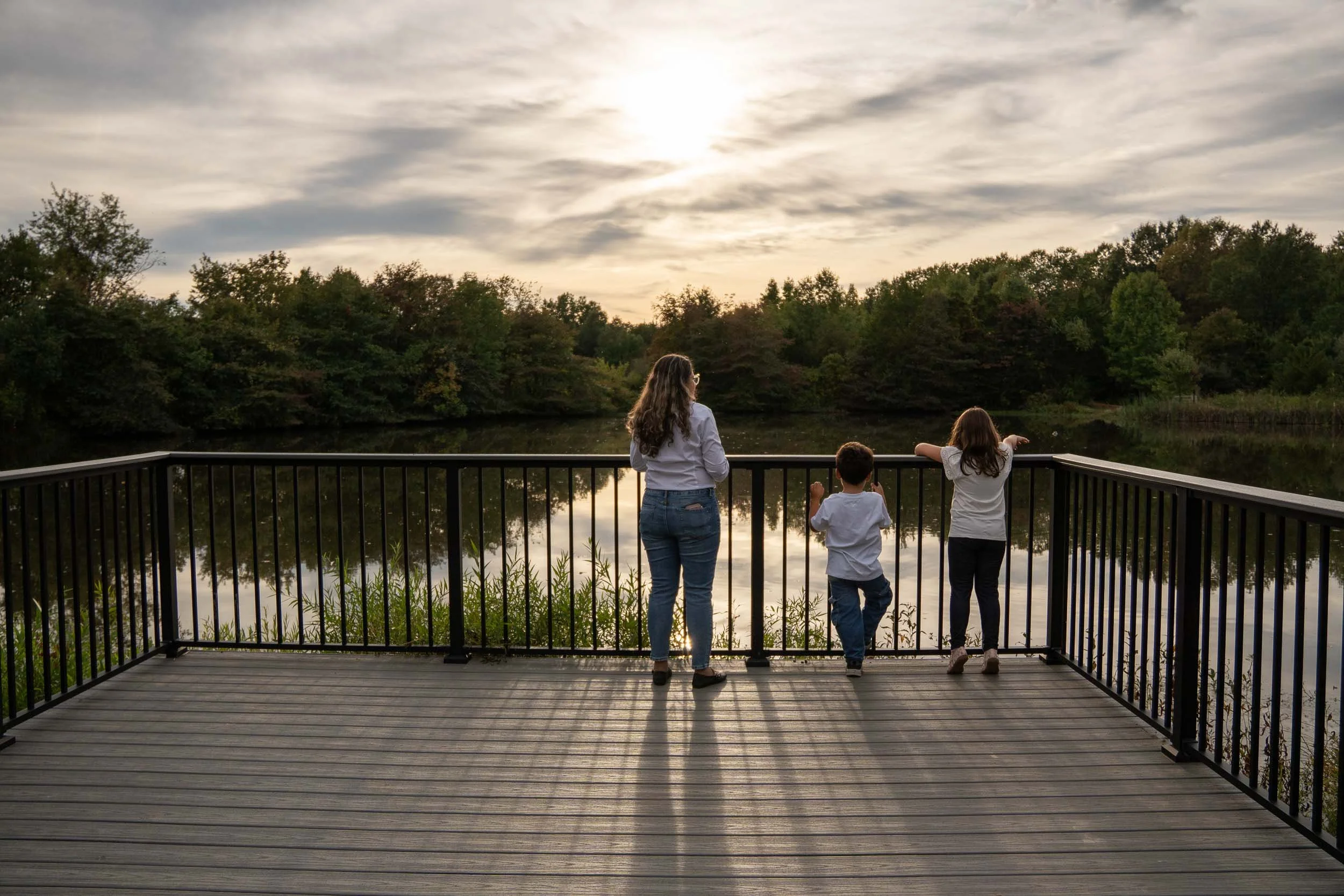Mother and two children standing at a lakeside overlook railing, looking out over the water at sunset.