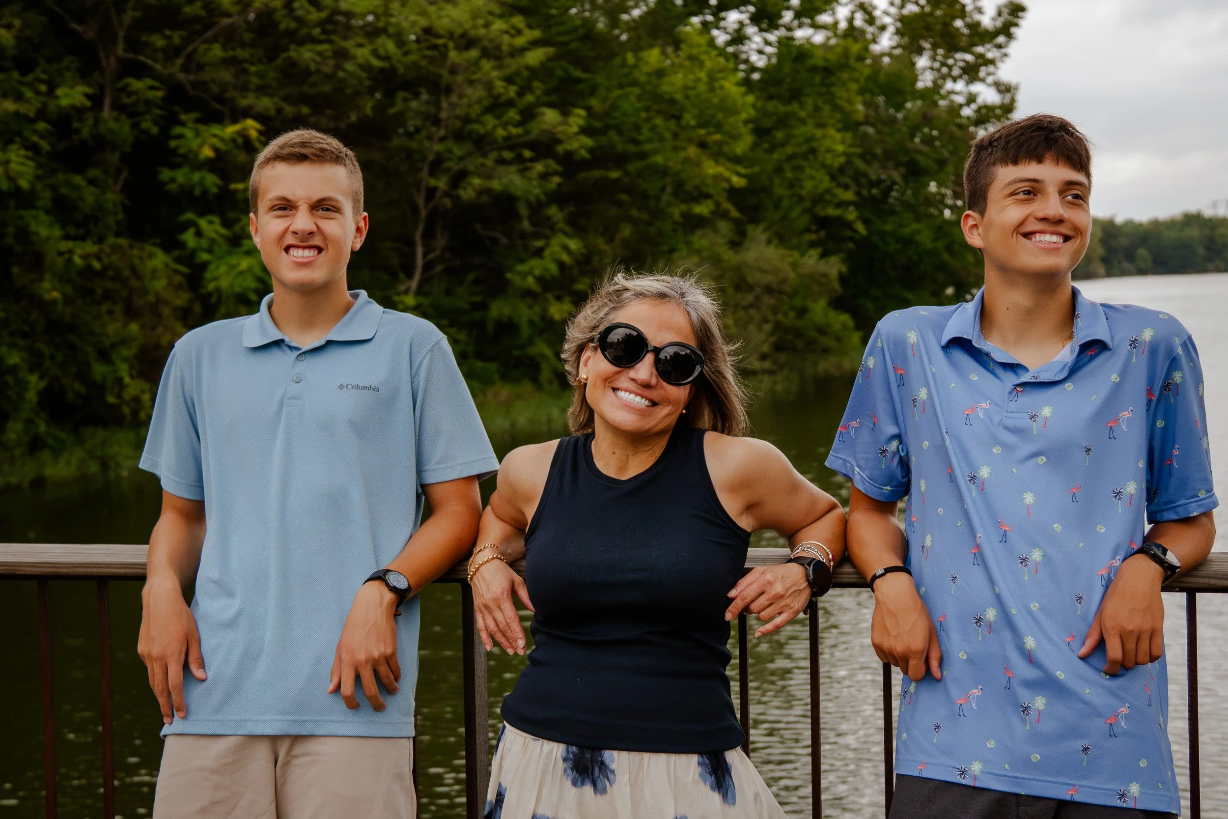 Group of three people standing outdoors near water, smiling and posing for a photo. The group includes two young men and one woman with sunglasses, all dressed casually, with trees in the background.