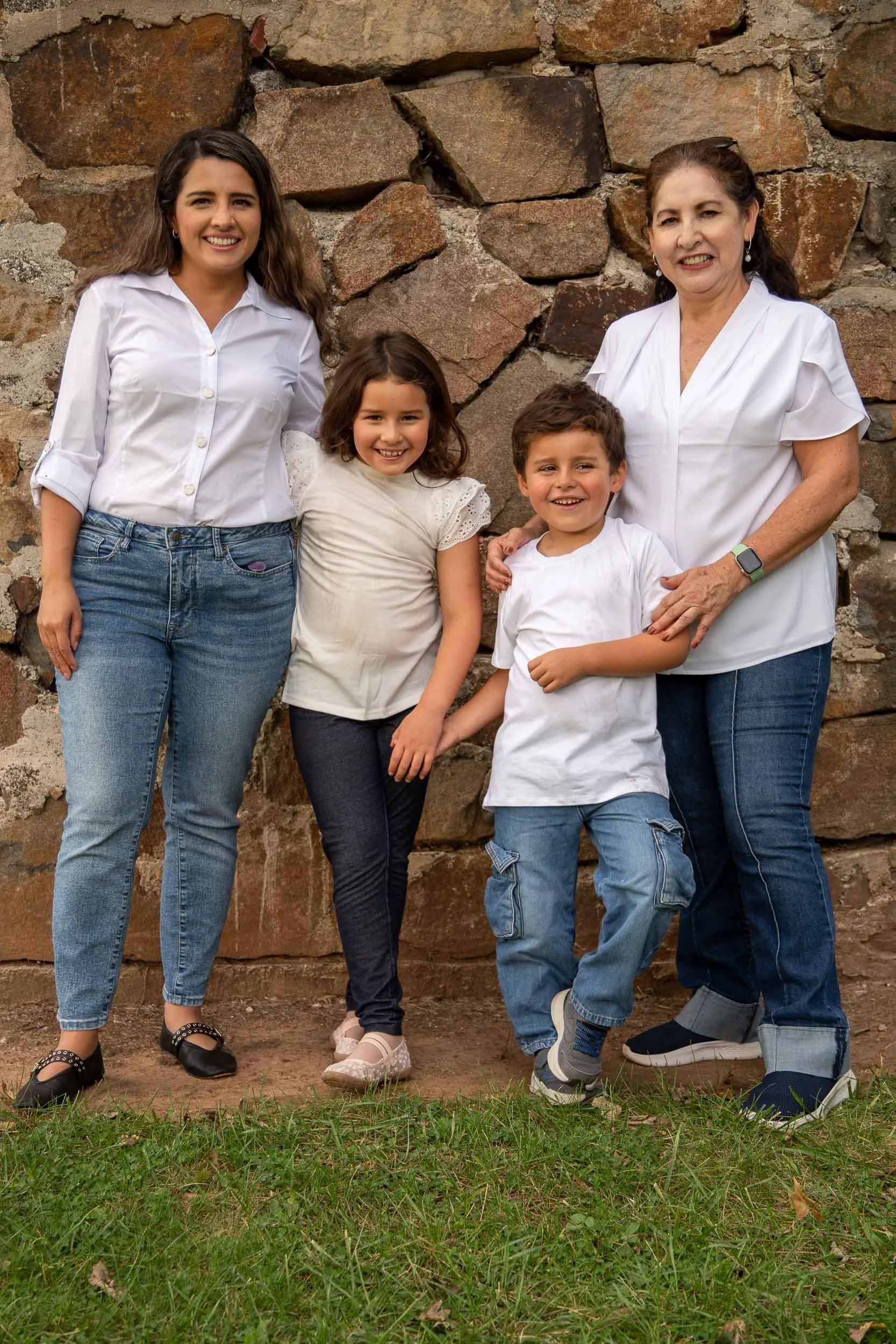 Grandmother, mother and two young children standing together and smiling in front of a fieldstone wall.