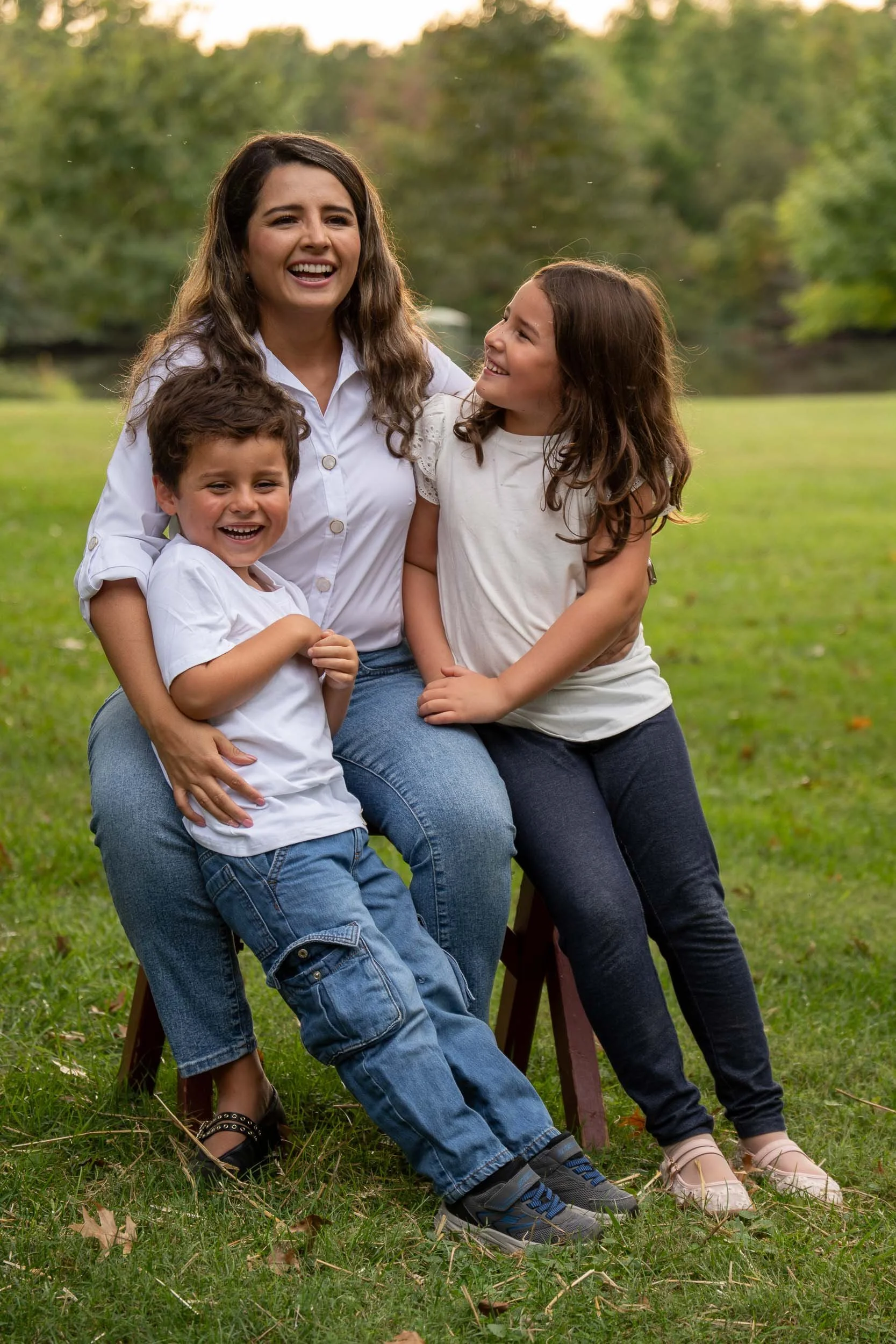 Mother laughing on a wooden stool in a park while her son and daughter lean into her.