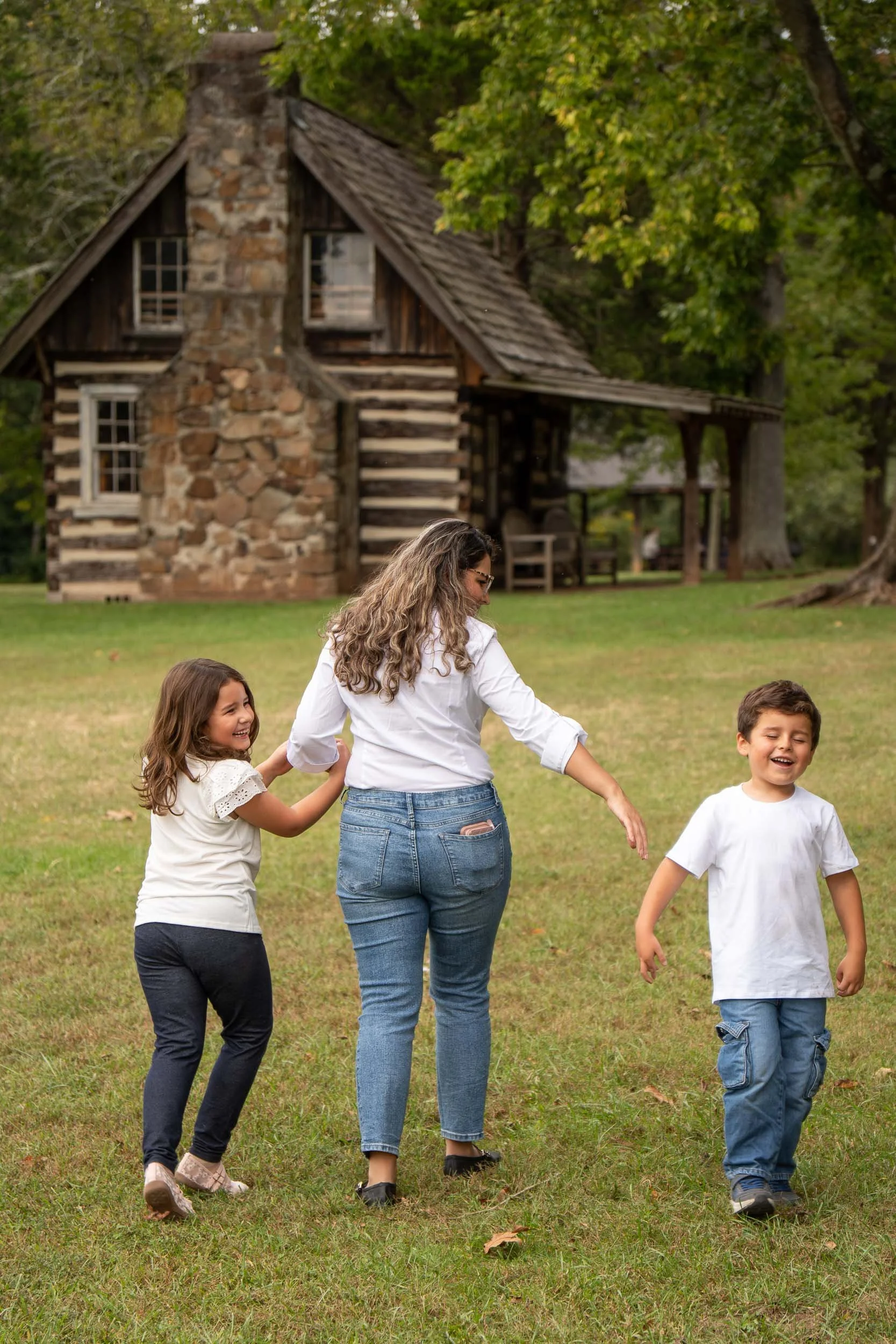 Mother walking with her son and daughter across a grassy field toward a historic log cabin with a stone chimney.
