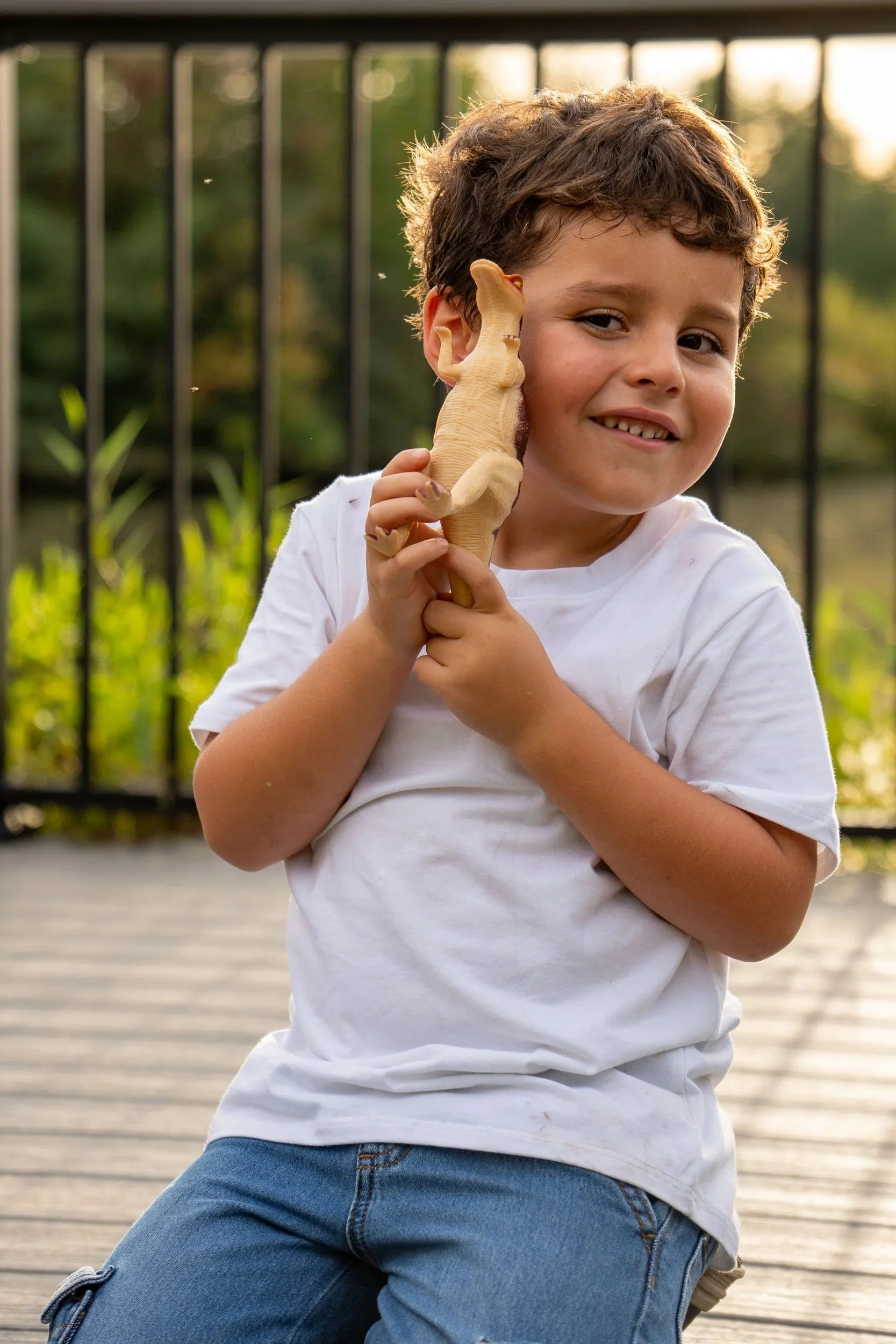 Smiling young boy holding a toy dinosaur up to his cheek on a lakeside boardwalk.