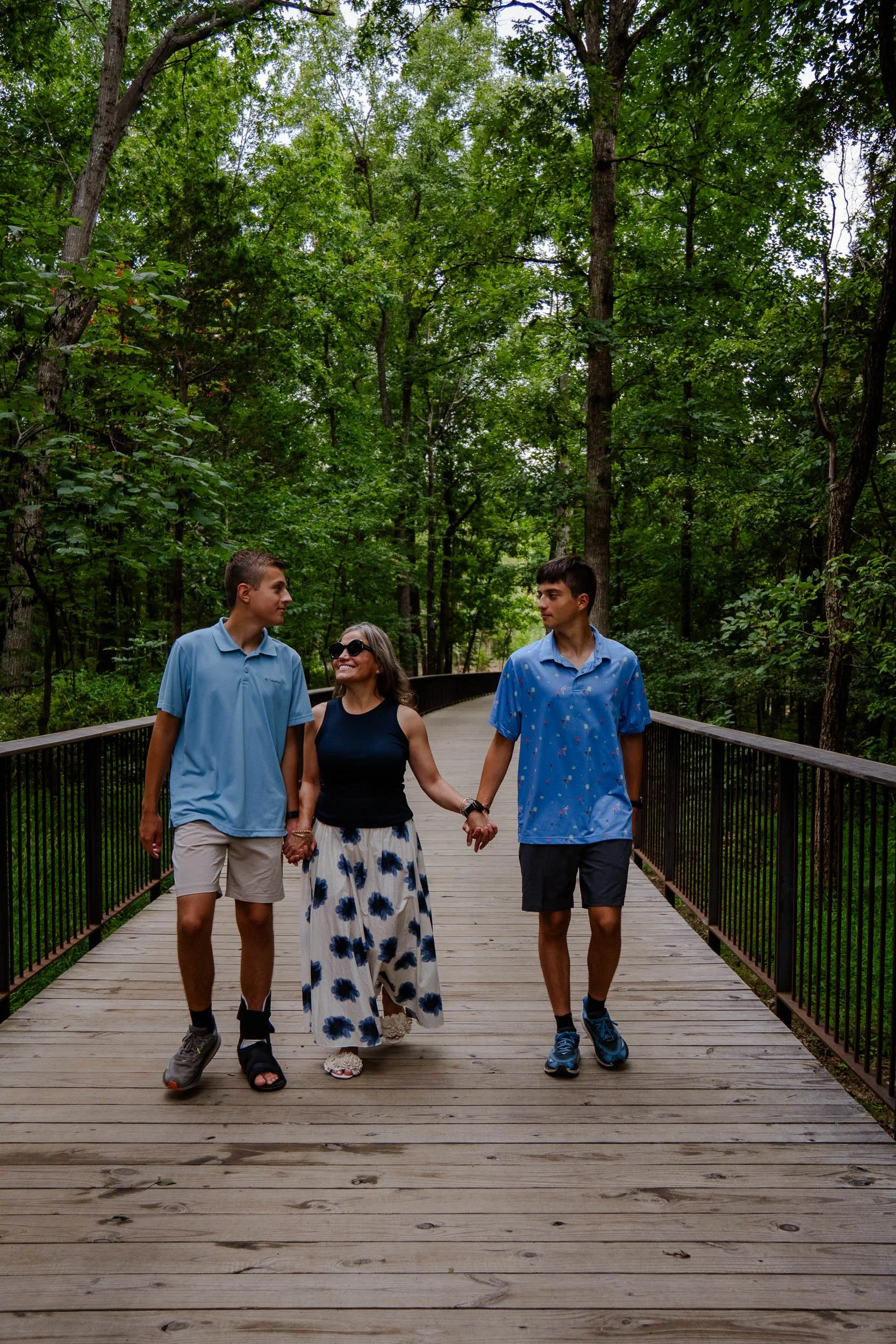 Three people walking on a wooden bridge in a forest, holding hands, smiling, and talking.