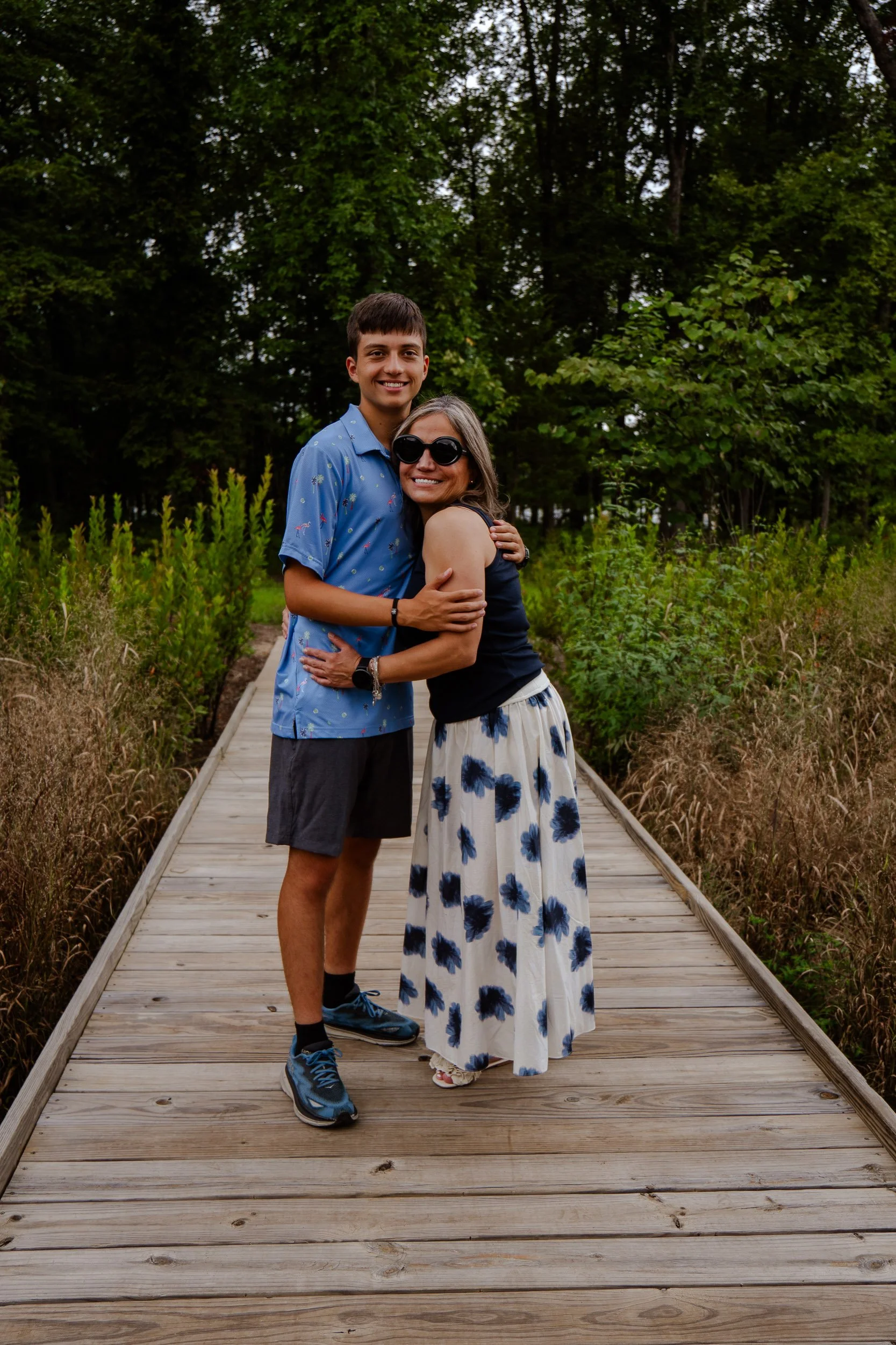 A young man and woman hugging on a wooden boardwalk surrounded by greenery and trees.