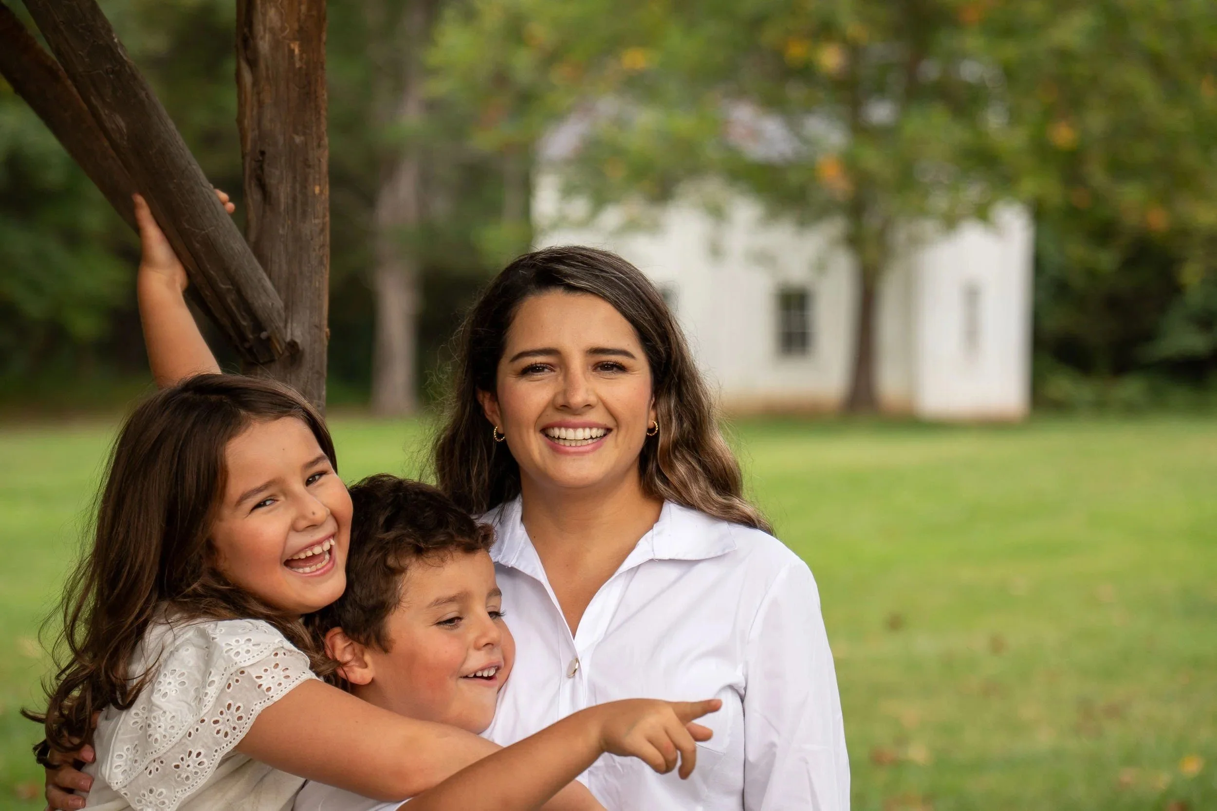 Mother laughing with her son and daughter, the girl pointing at something off camera, white farmhouse behind them.