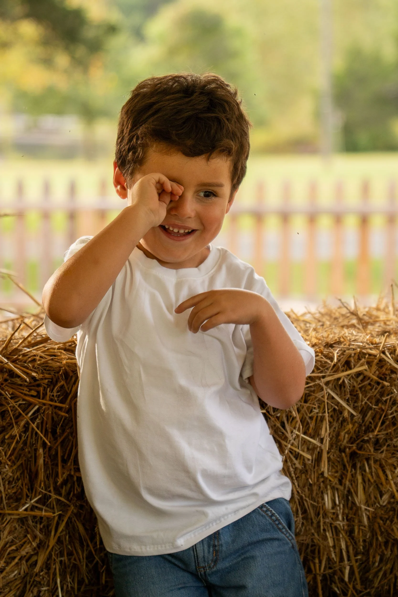 Young boy grinning shyly and rubbing one eye in front of hay bales and a white picket fence.
