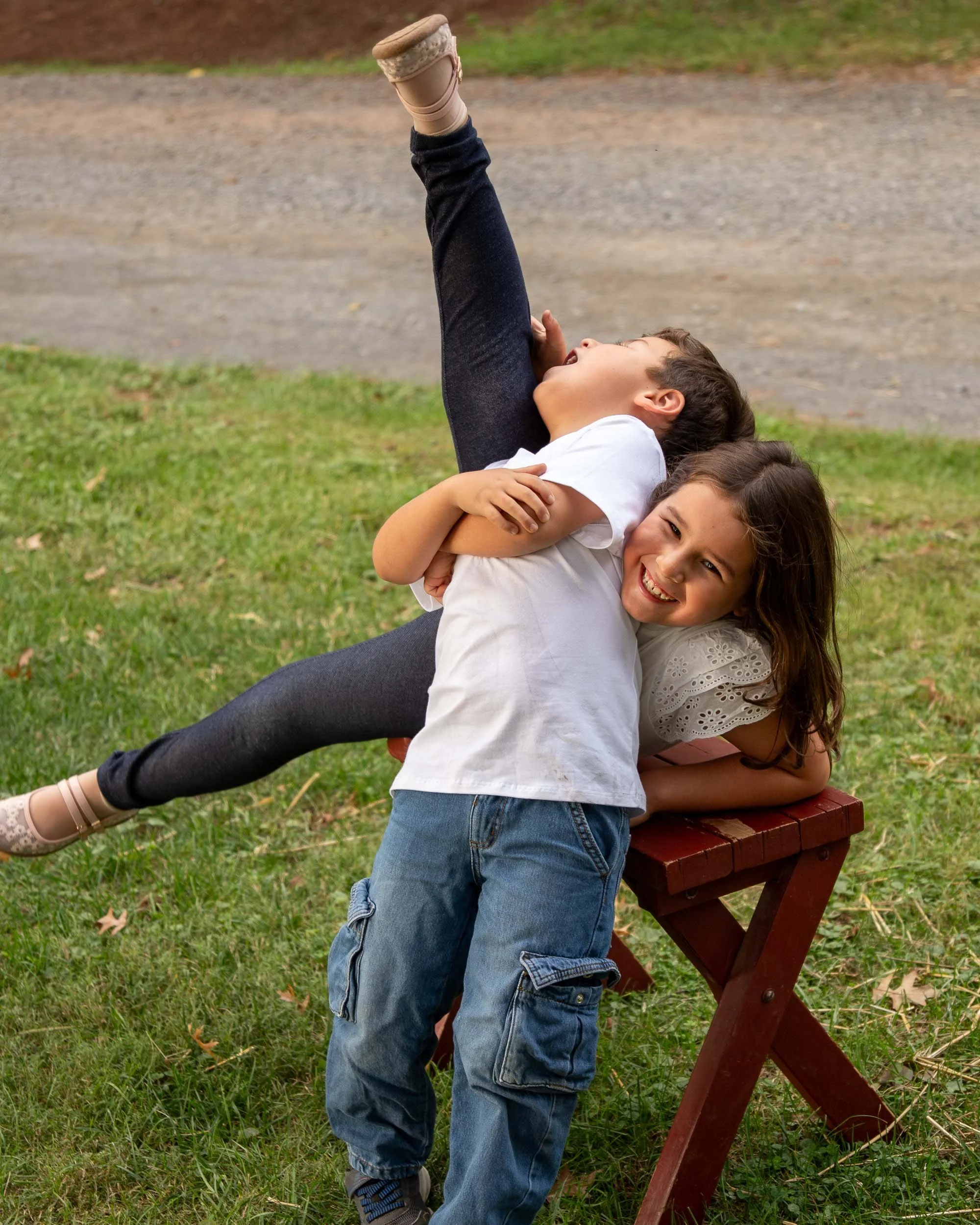A girl kicks one leg high in the air while her brother holds on to her and laughs, on a red wooden bench in the grass.