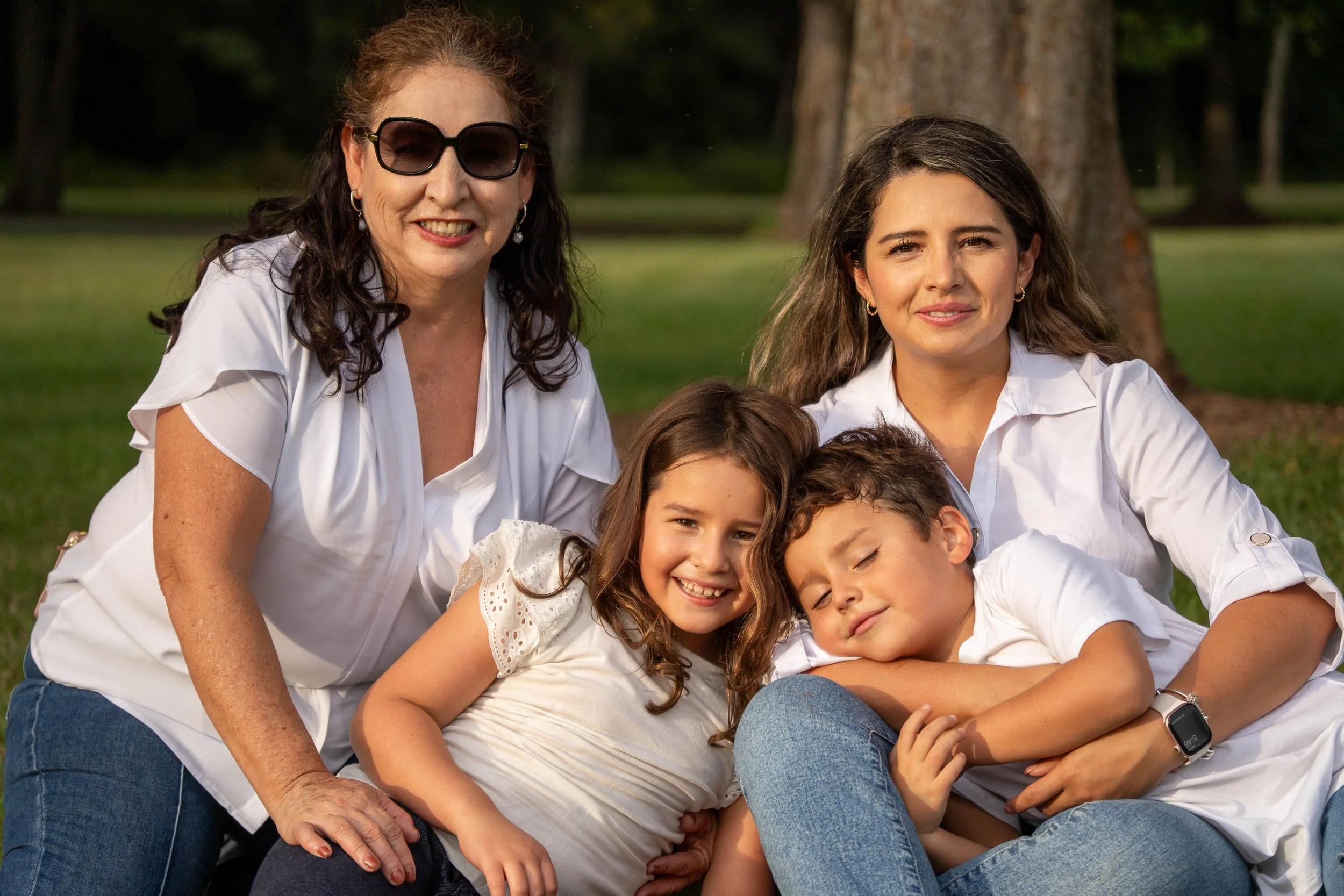 Grandmother and mother sitting in the grass with two children curled up in their laps, the boy resting with his eyes closed.