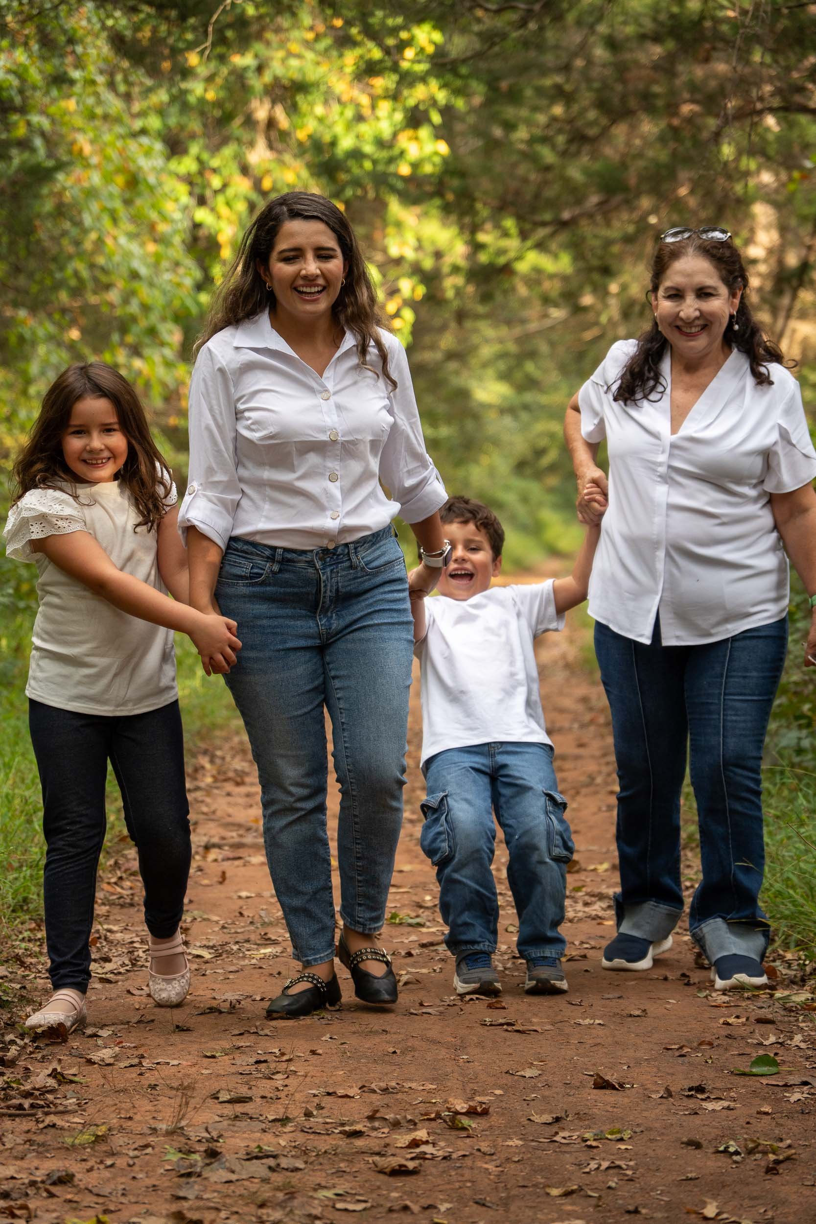 Grandmother, mother and two young children holding hands and laughing as they walk toward the camera on a wooded dirt trail.