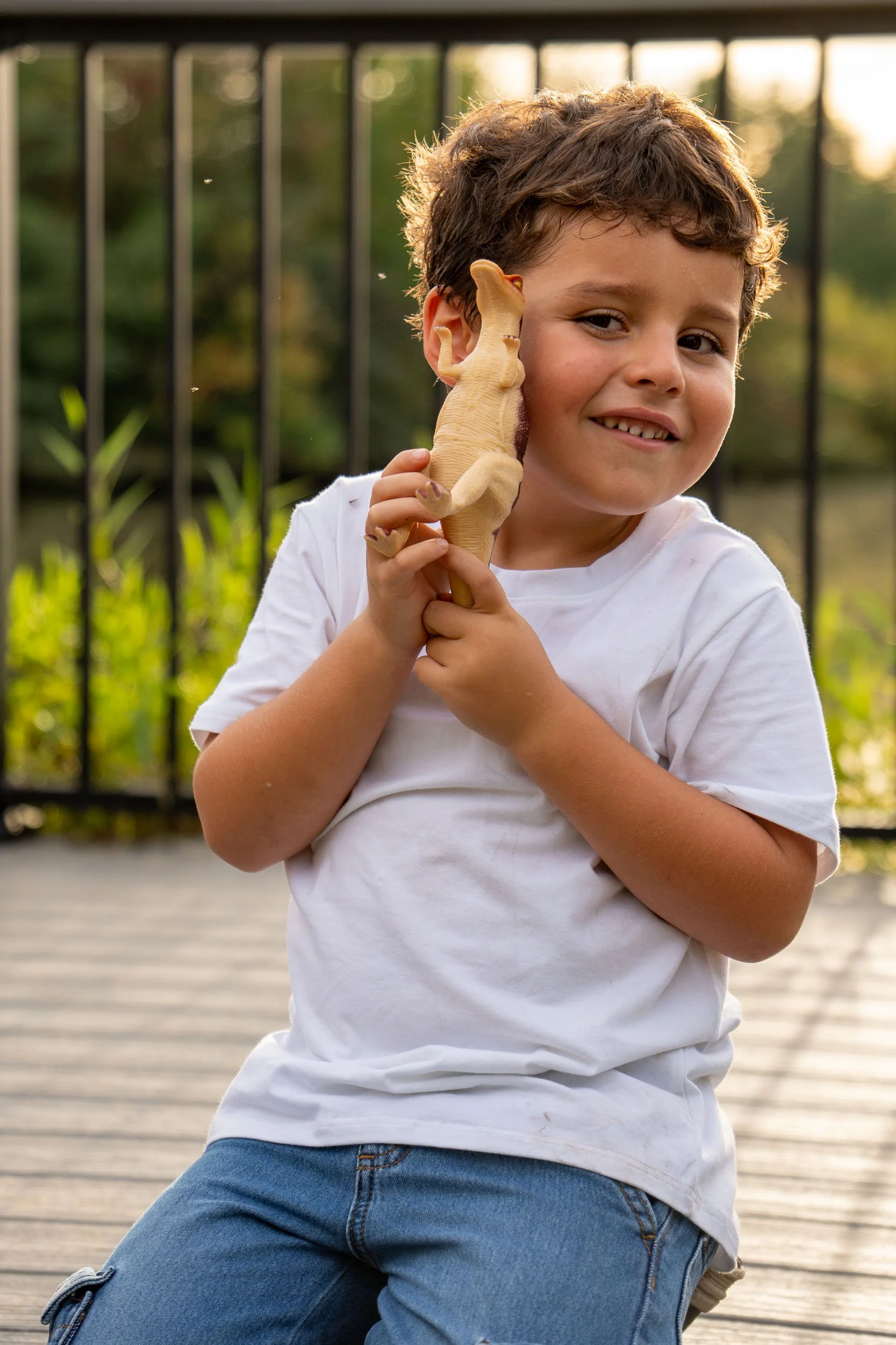 Smiling young boy holding a toy dinosaur up to his cheek on a lakeside boardwalk.