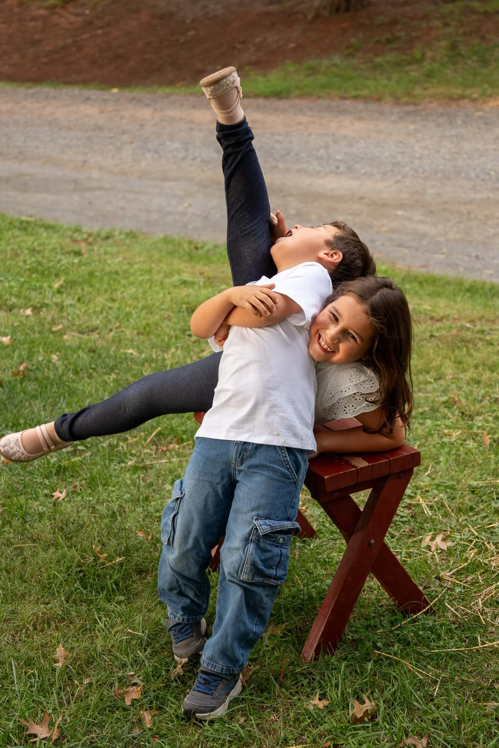 Young boy leaning back laughing as his sister kicks one leg high beside a small wooden bench in the grass.
