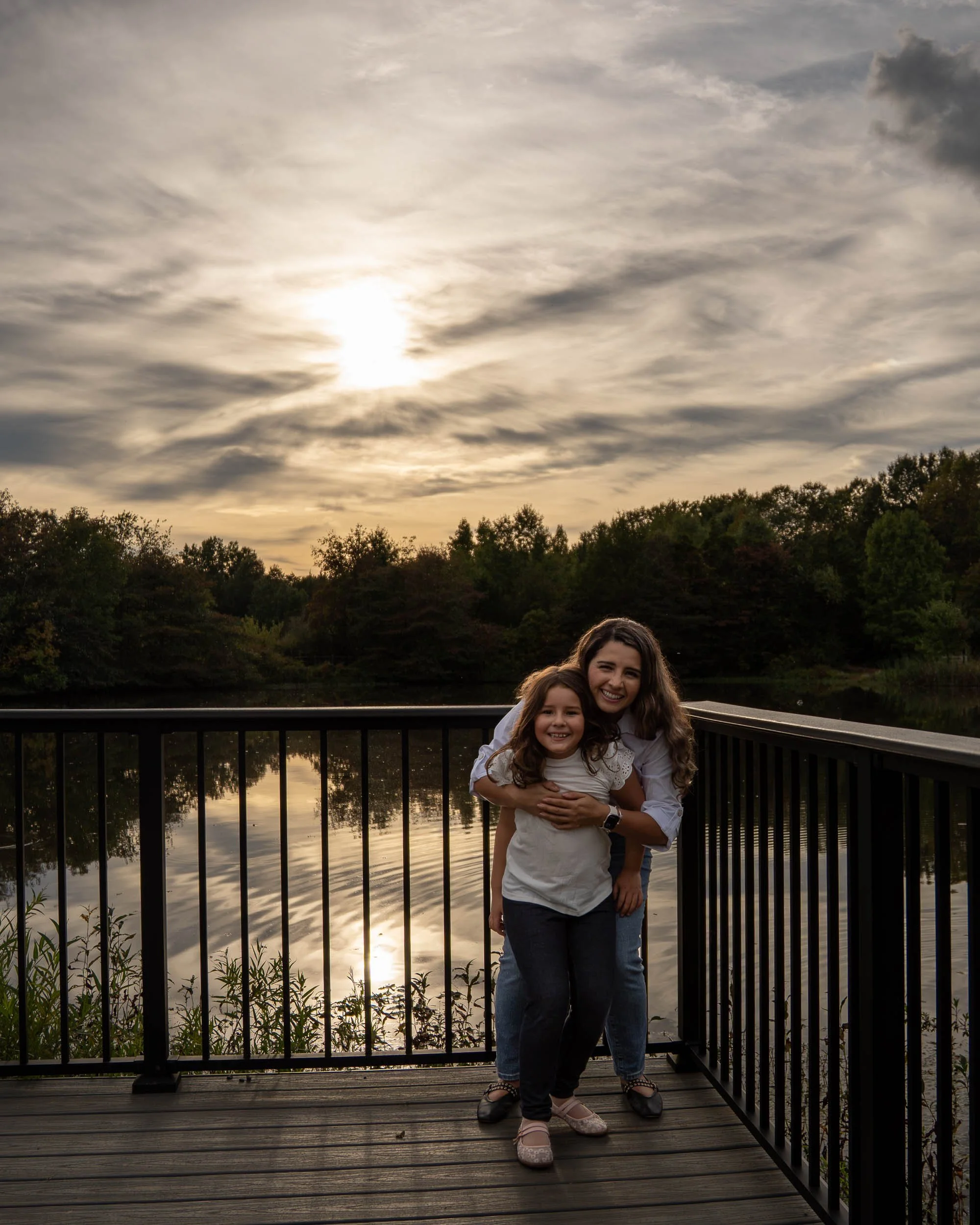 A mom hugs her daughter on a lakeside dock at sunset.