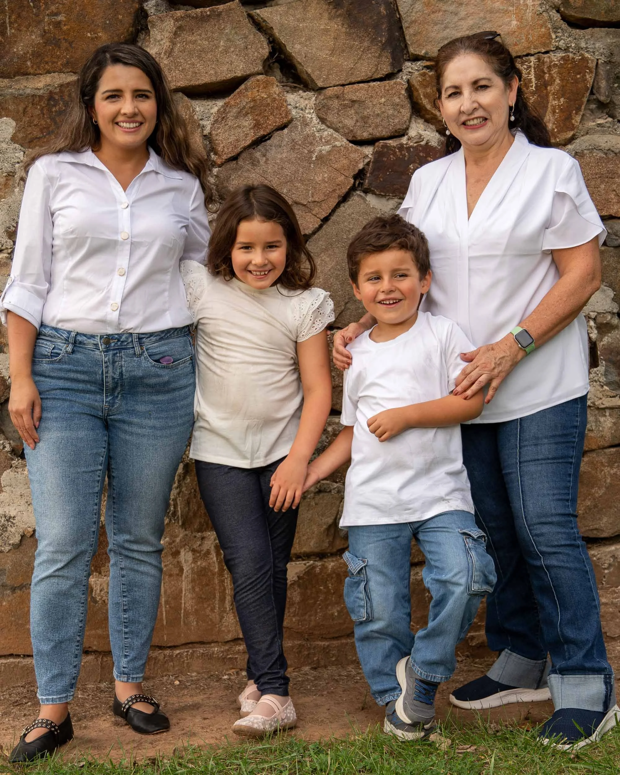 A mom, grandmother and two kids stand together in front of an old stone wall, smiling at the camera.