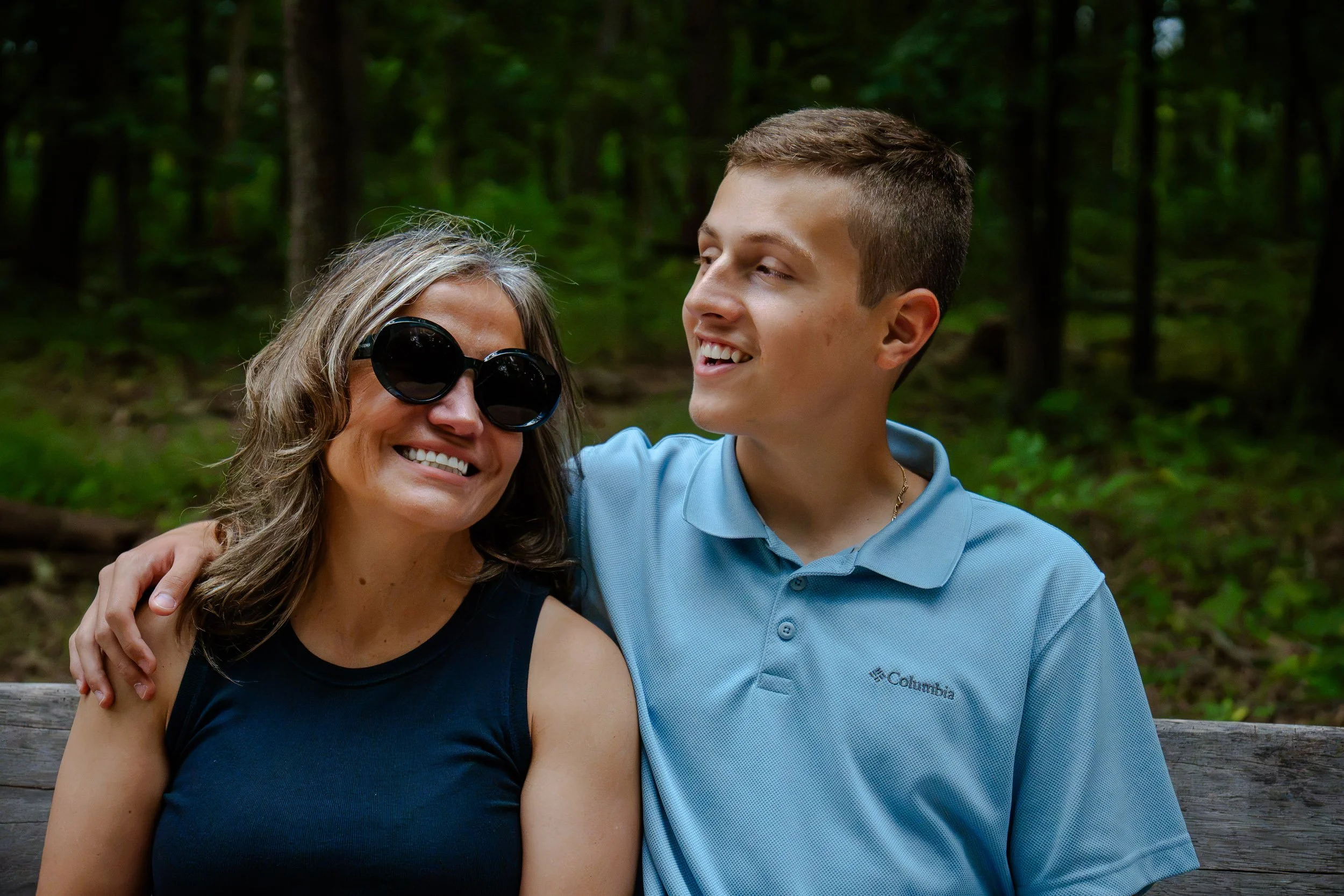 A woman with gray hair and sunglasses smiling, sitting next to a young man wearing a blue polo shirt, both smiling and looking happy, outdoors in a forested area.