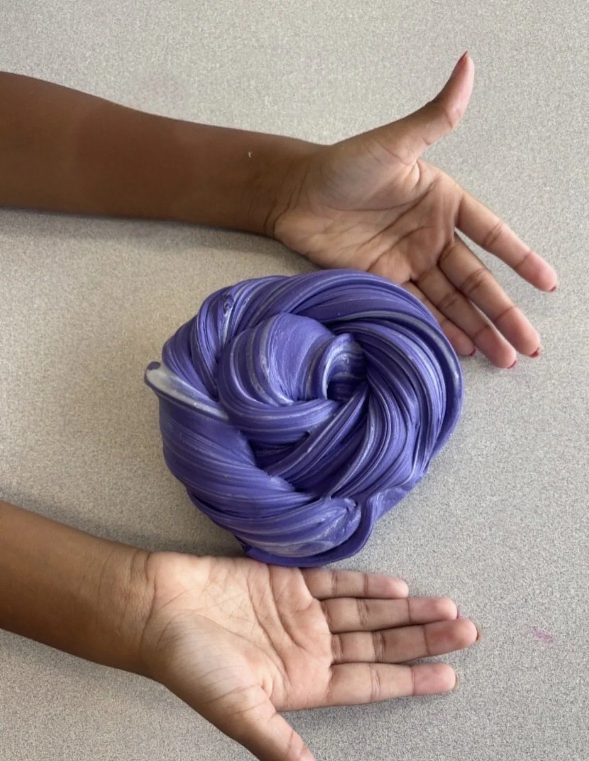 Two hands with brown skin, one above and one below, holding a twisted ball of purple and white marble-like material on a light gray surface.