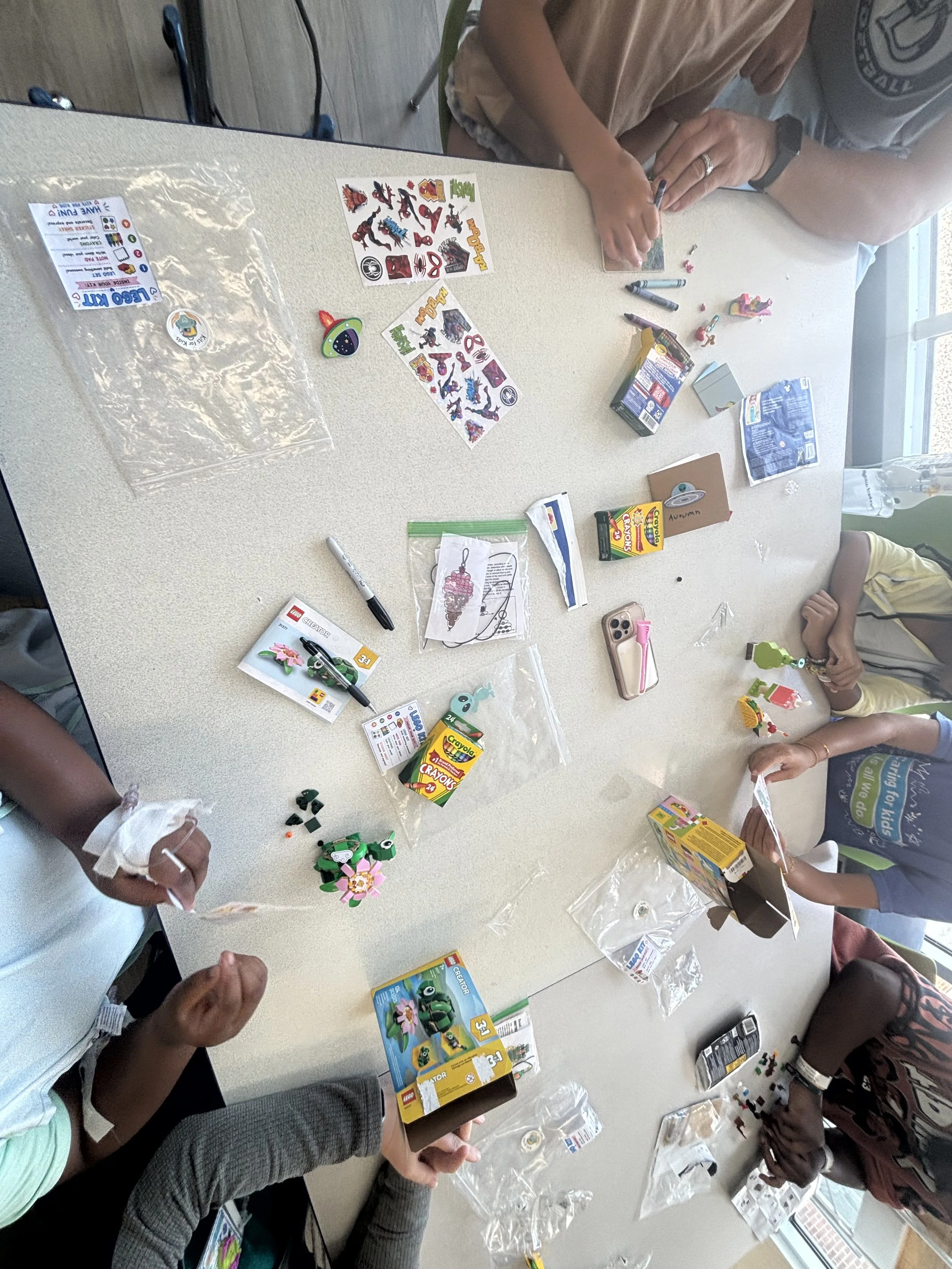 People assembling colorful LEGO sets on a table with various LEGO boxes, instructions, and pieces scattered around.