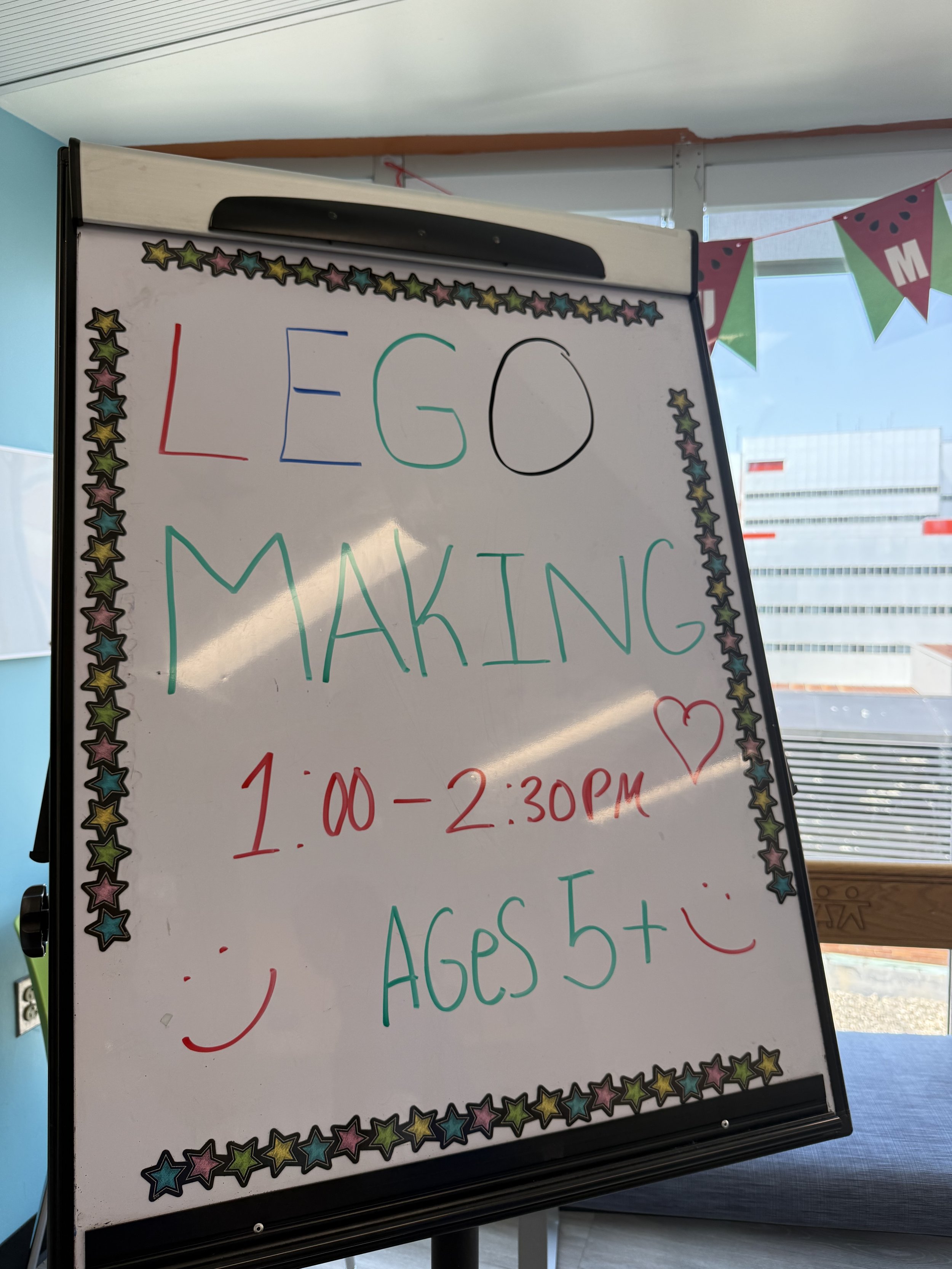 Whiteboard with text about Lego making event, decorated with colorful star border, in a room with windows and a red and green pennant banner.