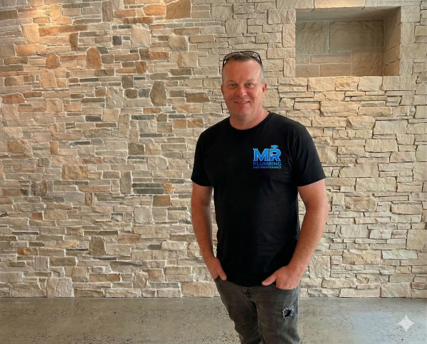 A smiling man in a black t-shirt with blue text stands against a beige stone wall, with sunglasses resting on his head and his hands in his pockets.