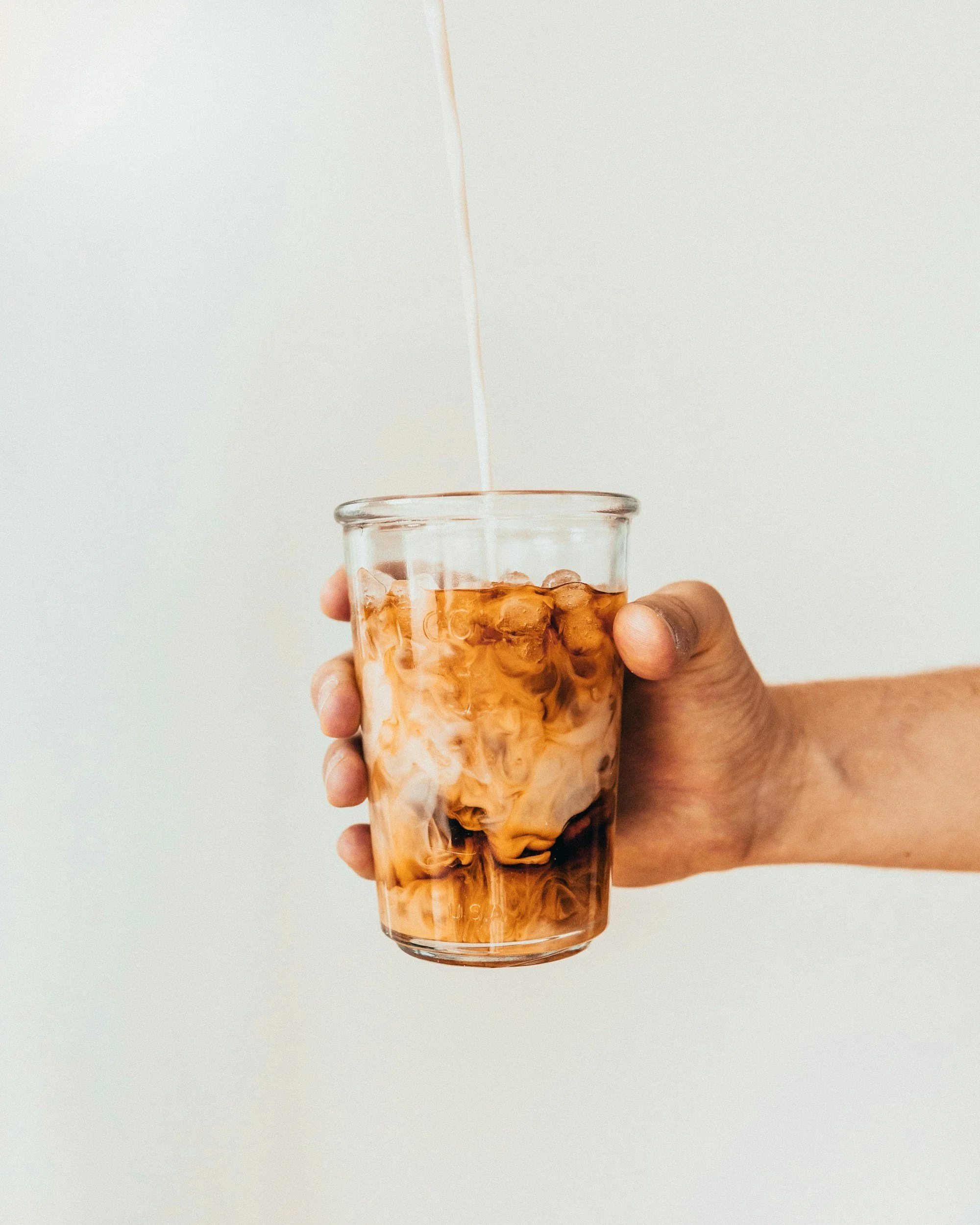A hand holding a glass of iced tea with ice cubes, with a stream of pouring tea above the glass.