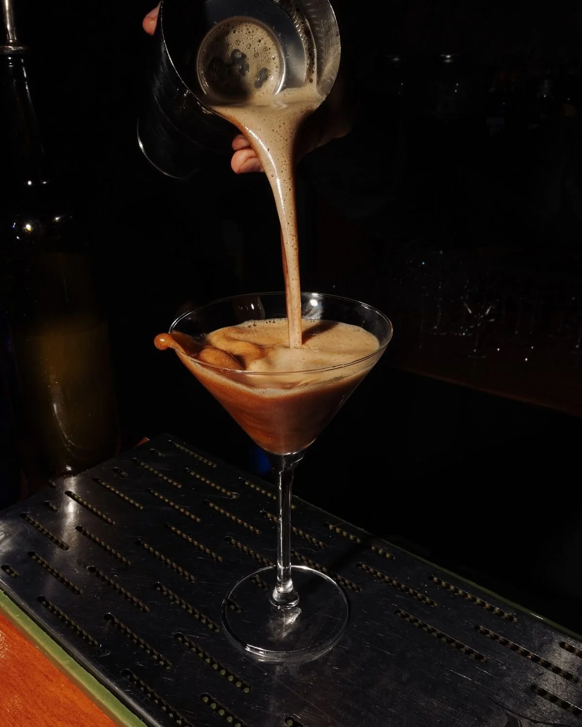 A bartender pours a creamy, frothy coffee cocktail into a martini glass in a dark bar setting.