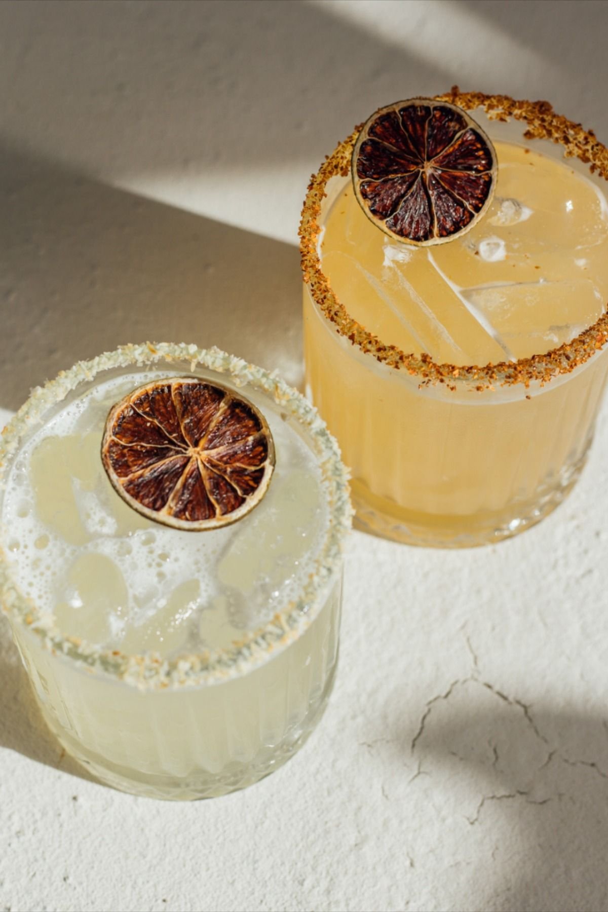 Two glasses of a yellow beverage garnished with dried citrus slices, with salted rims and ice. The background is a light, textured surface.