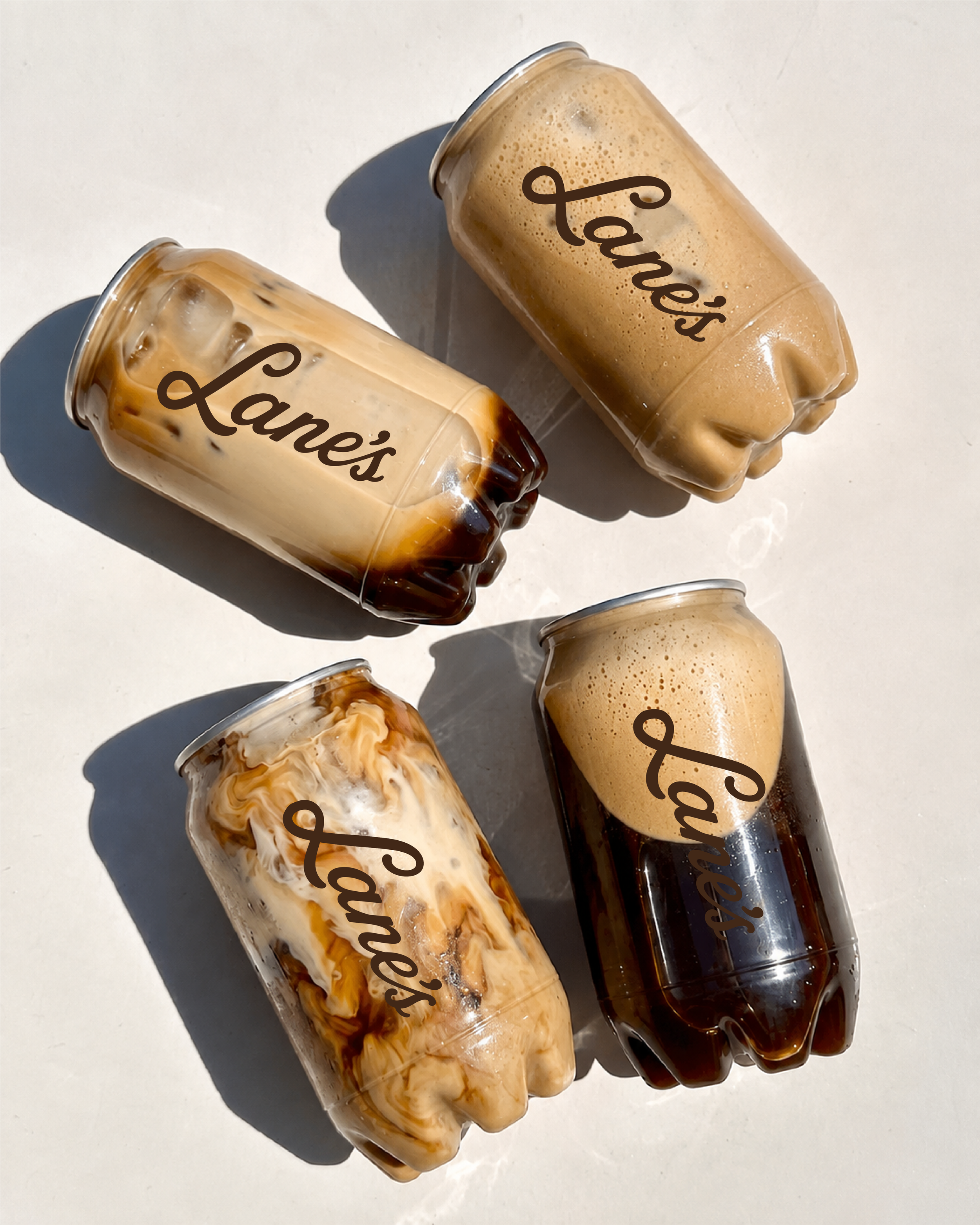 Three iced coffee drinks in clear plastic cups with 'Lane's' written on them, placed on a white surface in sunlight, casting shadows.