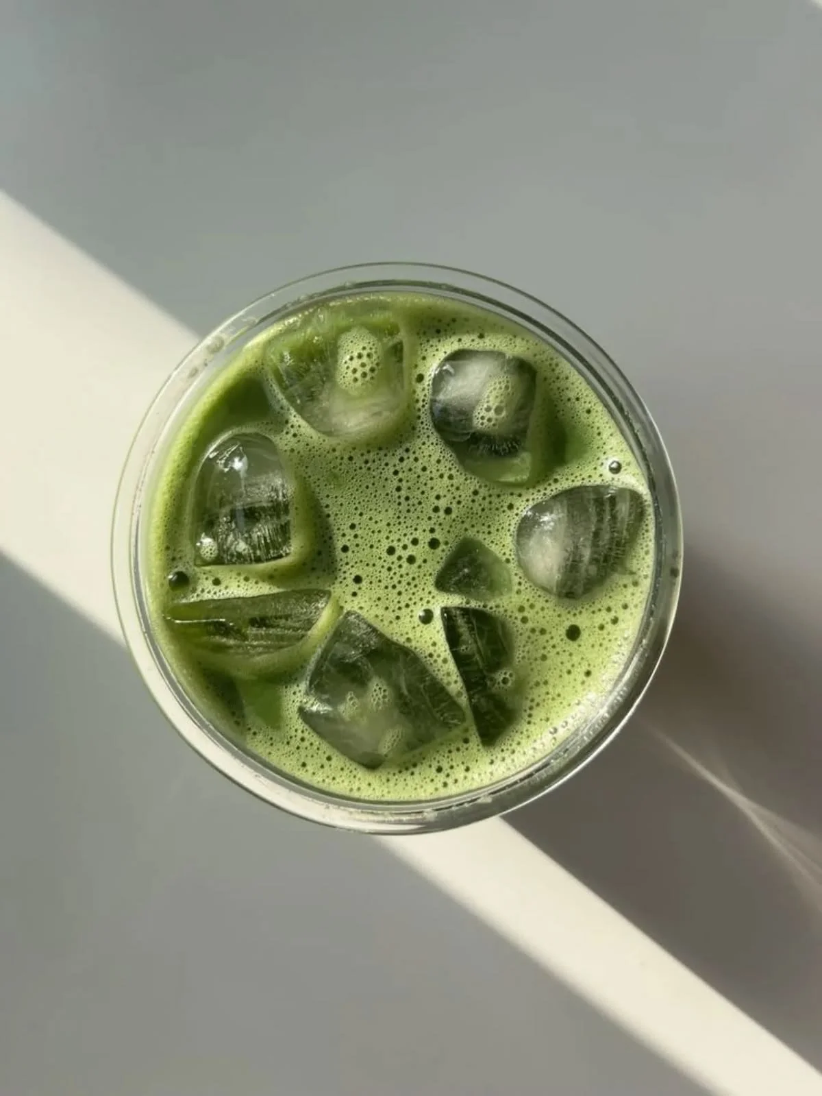 A glass of iced matcha latte with green tea and ice cubes, viewed from above on a white and gray surface.