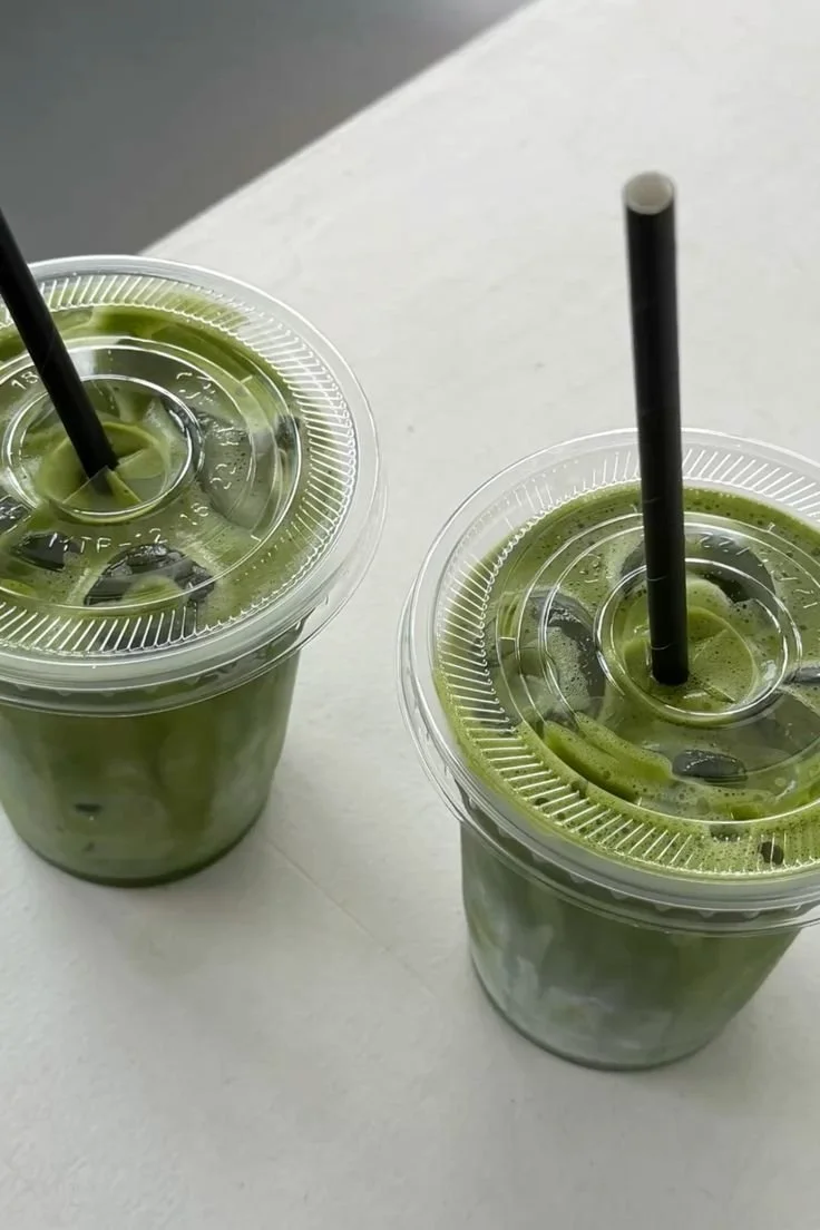 Two plastic cups with green beverages and black straws on a white surface.