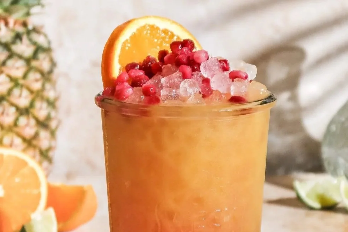 A glass of tropical fruit smoothie topped with pomegranate seeds, an orange slice, and crushed ice, with a pineapple and citrus fruits in the background.