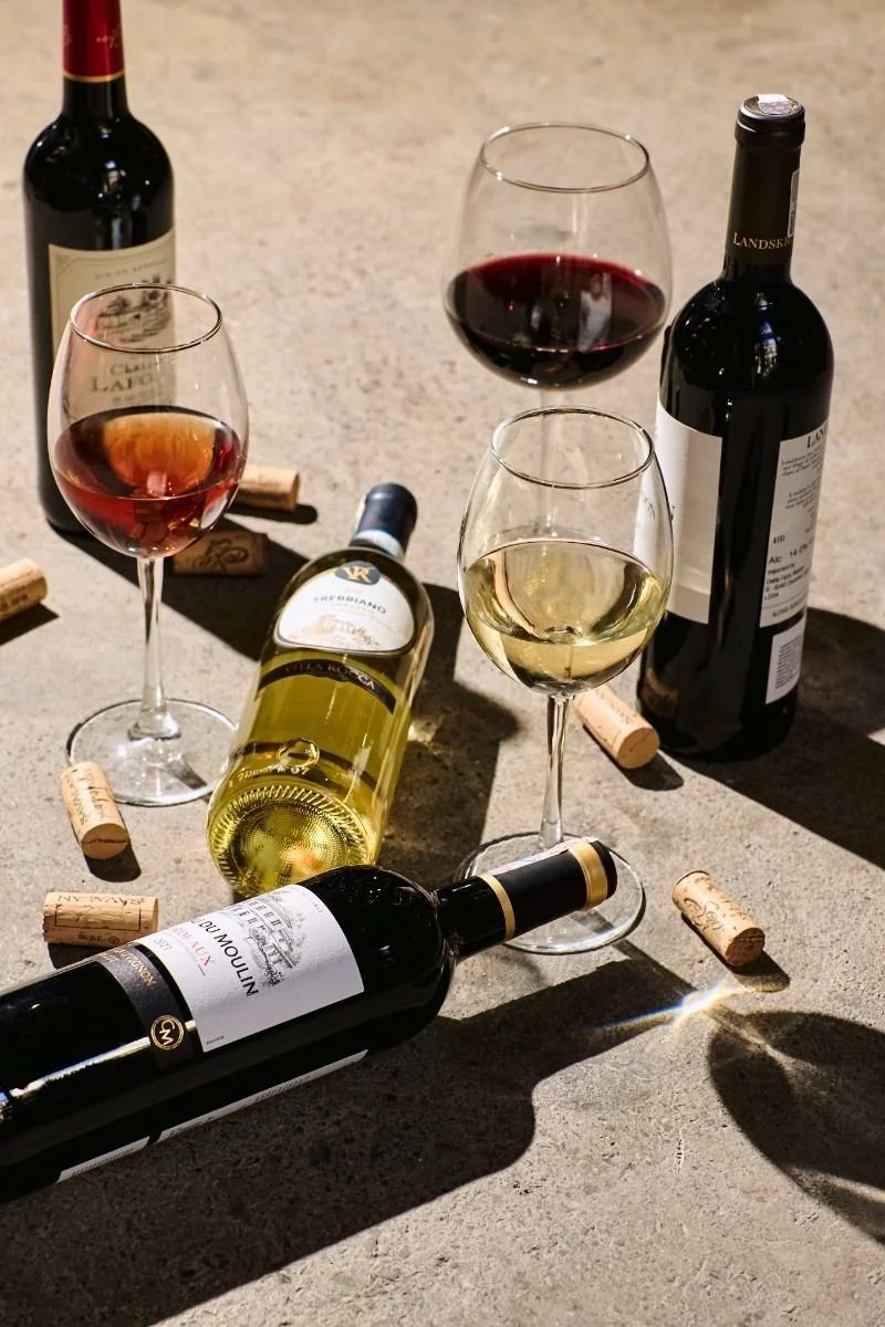 Various bottles of wine, corks, and wine glasses filled with red and white wine on a textured surface.