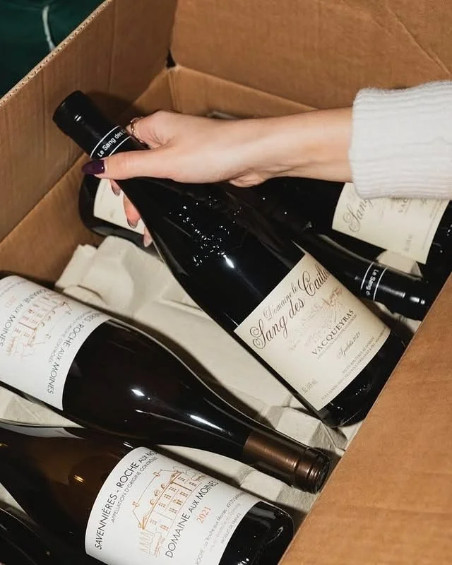 A person with light skin and purple nail polish holding a bottle of Sancerre wine above other wine bottles in a cardboard box.
