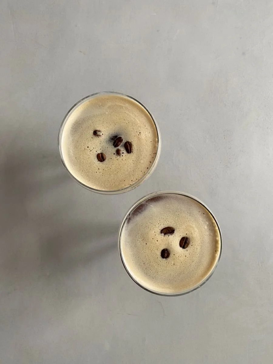 Two glasses of creamy coffee topped with coffee beans on a light gray surface.