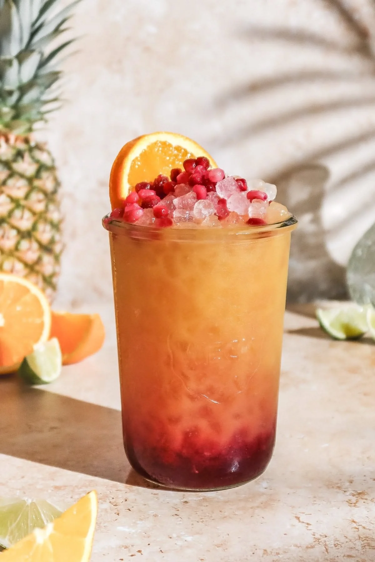 A vibrant, layered cocktail in a tall glass garnished with an orange slice and pink pomegranate seeds, with a tropical pineapple in the background.