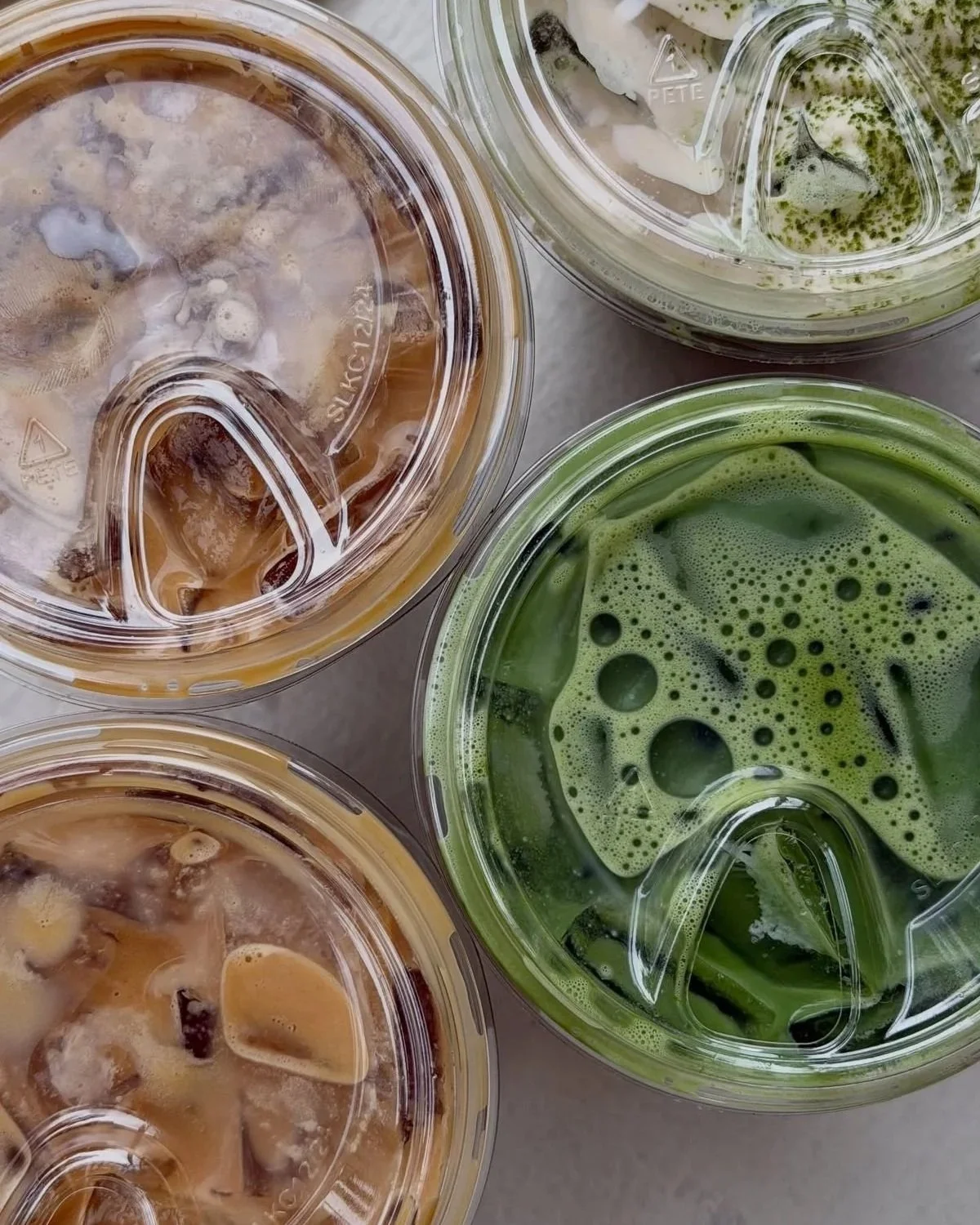 Multiple cups of iced coffee and matcha drinks in clear plastic cups, showing different shades of brown and green with ice cubes and foam.