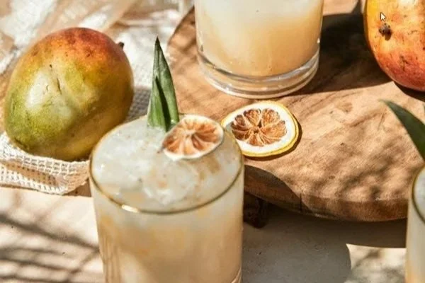 Two glasses of a creamy yellow beverage garnished with dried citrus slices, surrounded by whole mangos, dried citrus slices, and green leaves on a wooden surface.