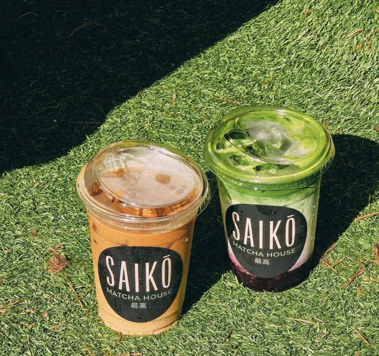 Two cups of matcha drinks from Saikō Matcha House on artificial grass with sunlight and shadows.