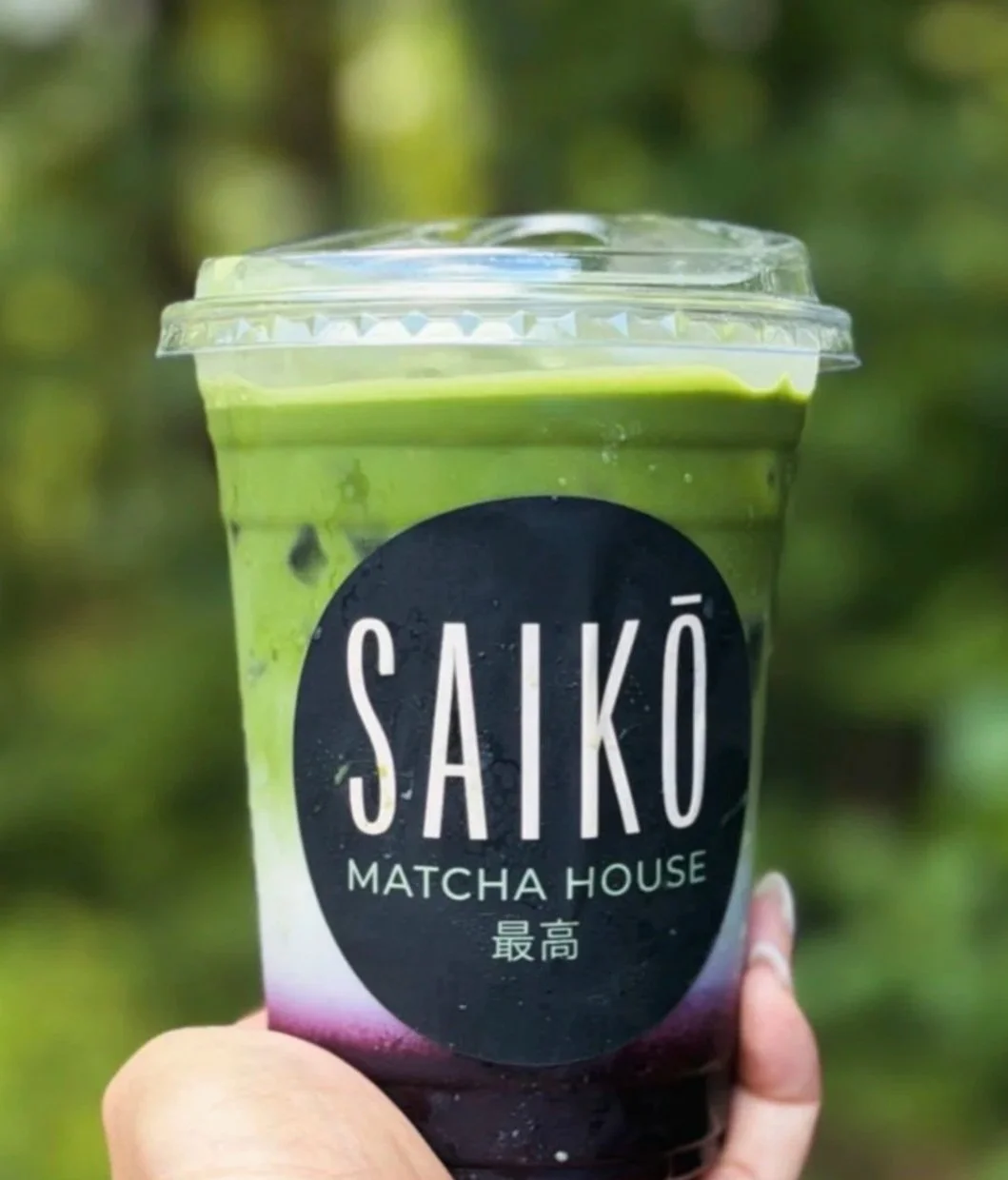 A plastic cup of matcha green tea latte with a lid, held outdoors with a green, blurred background.