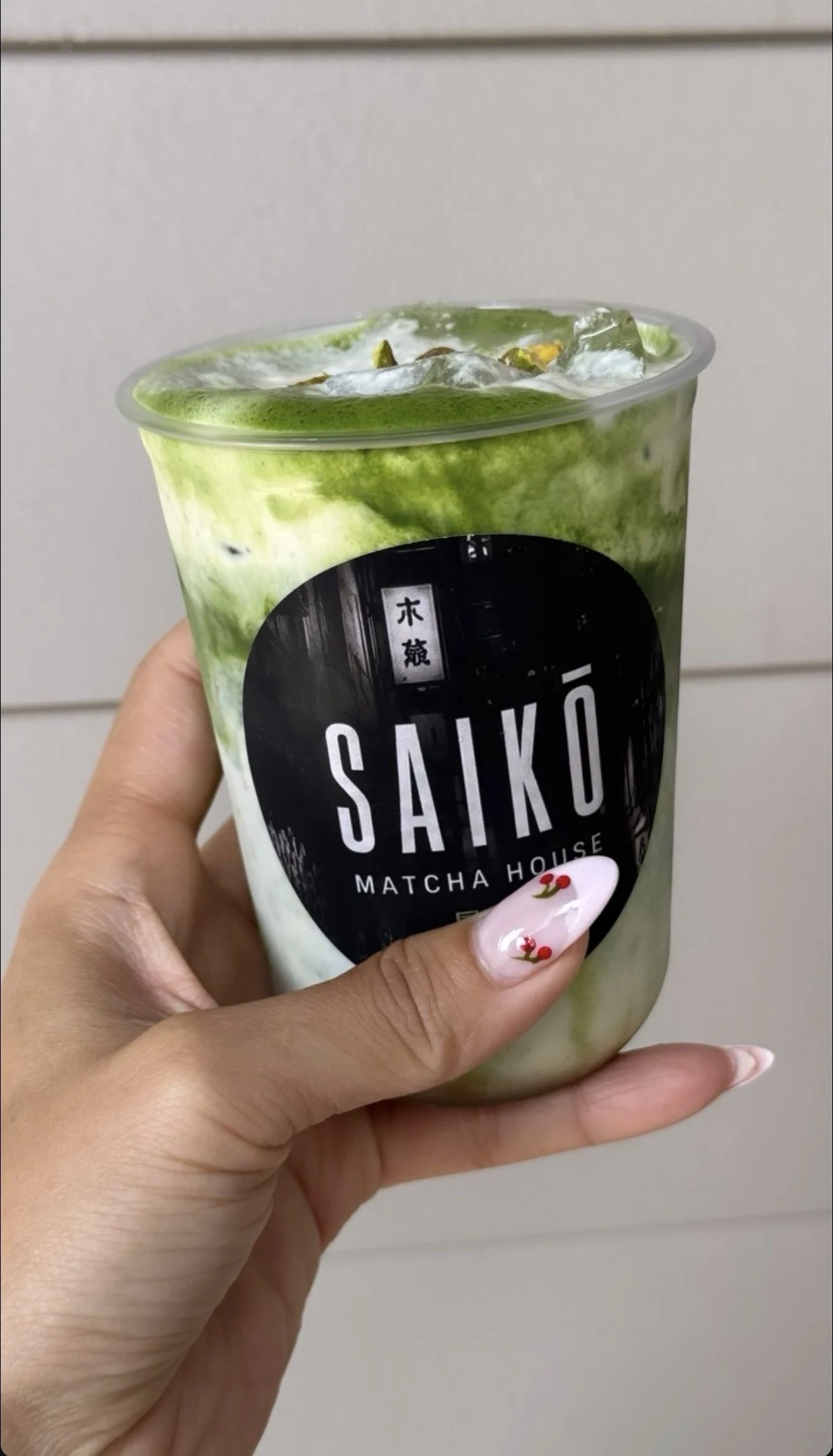 A hand holding a cup of matcha latte from Saiko Matcha House with ice and green matcha swirling in the cup.