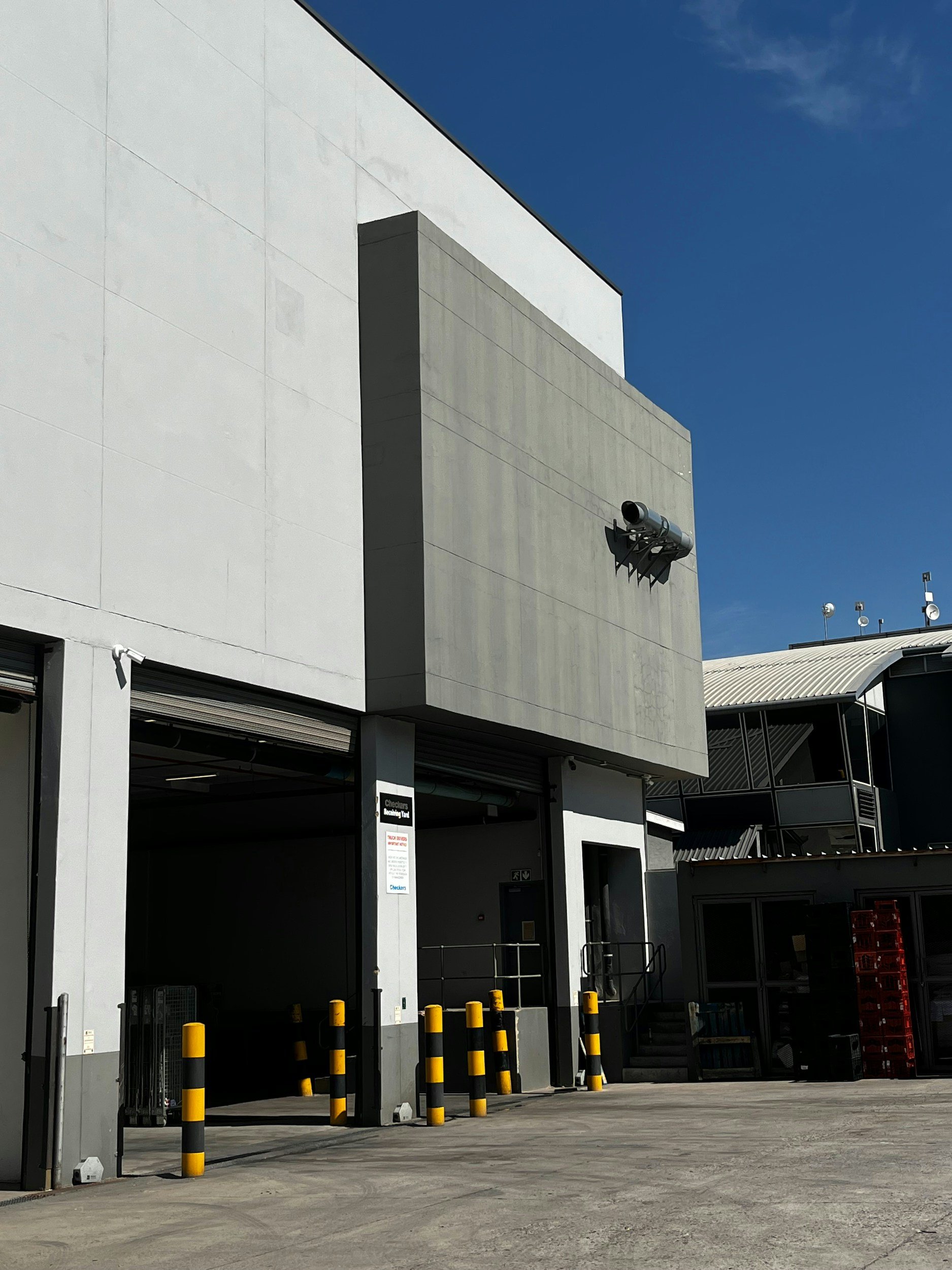 Exterior of a modern building with large gray and white walls, security cameras, and yellow-black striped bollards in the parking area. Clear blue sky in the background.