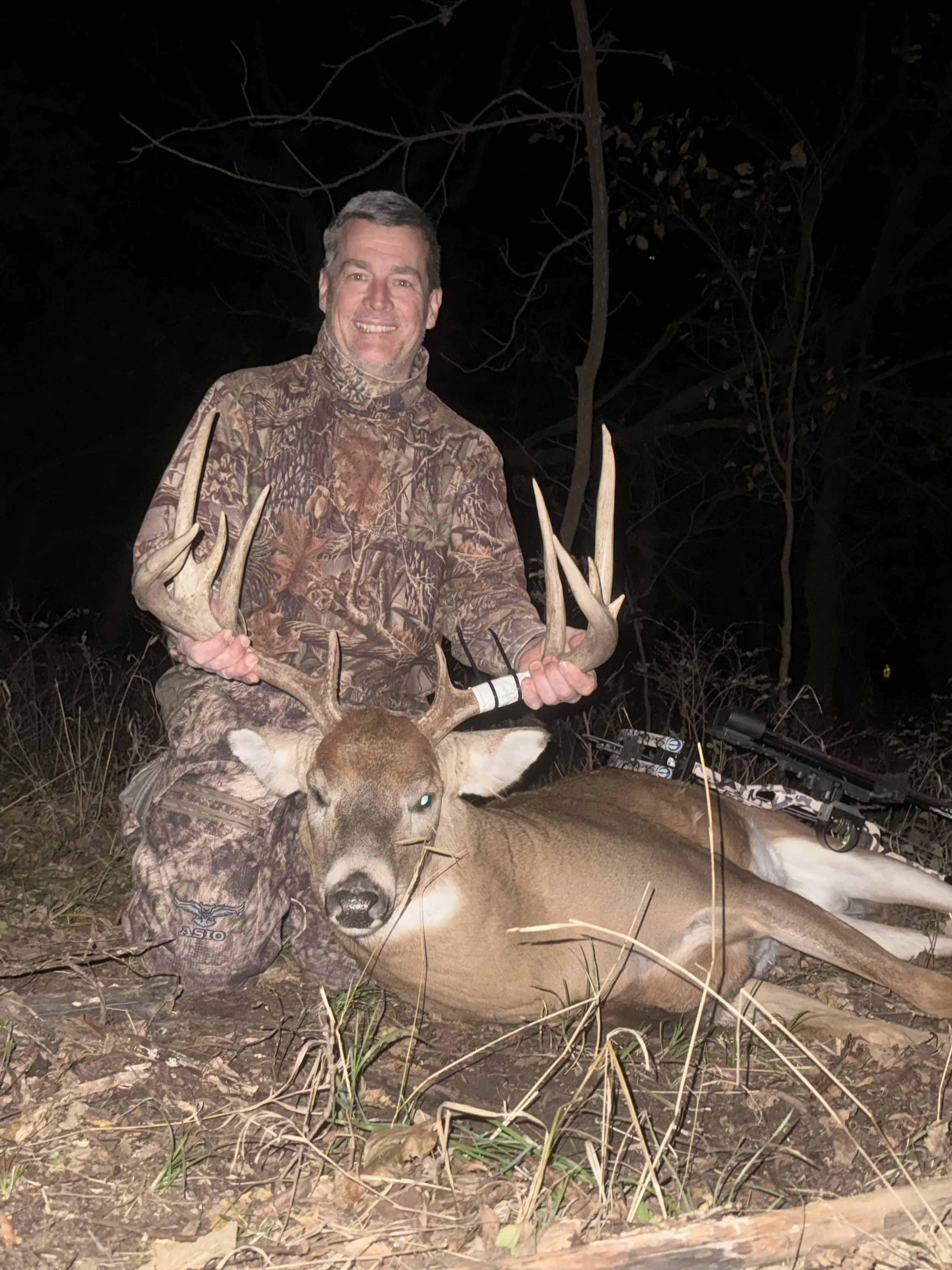 A smiling man dressed in camouflage clothing in a wooded area at night, holding the antlers of a large deer he has hunted, with a bow resting on the deer's back.