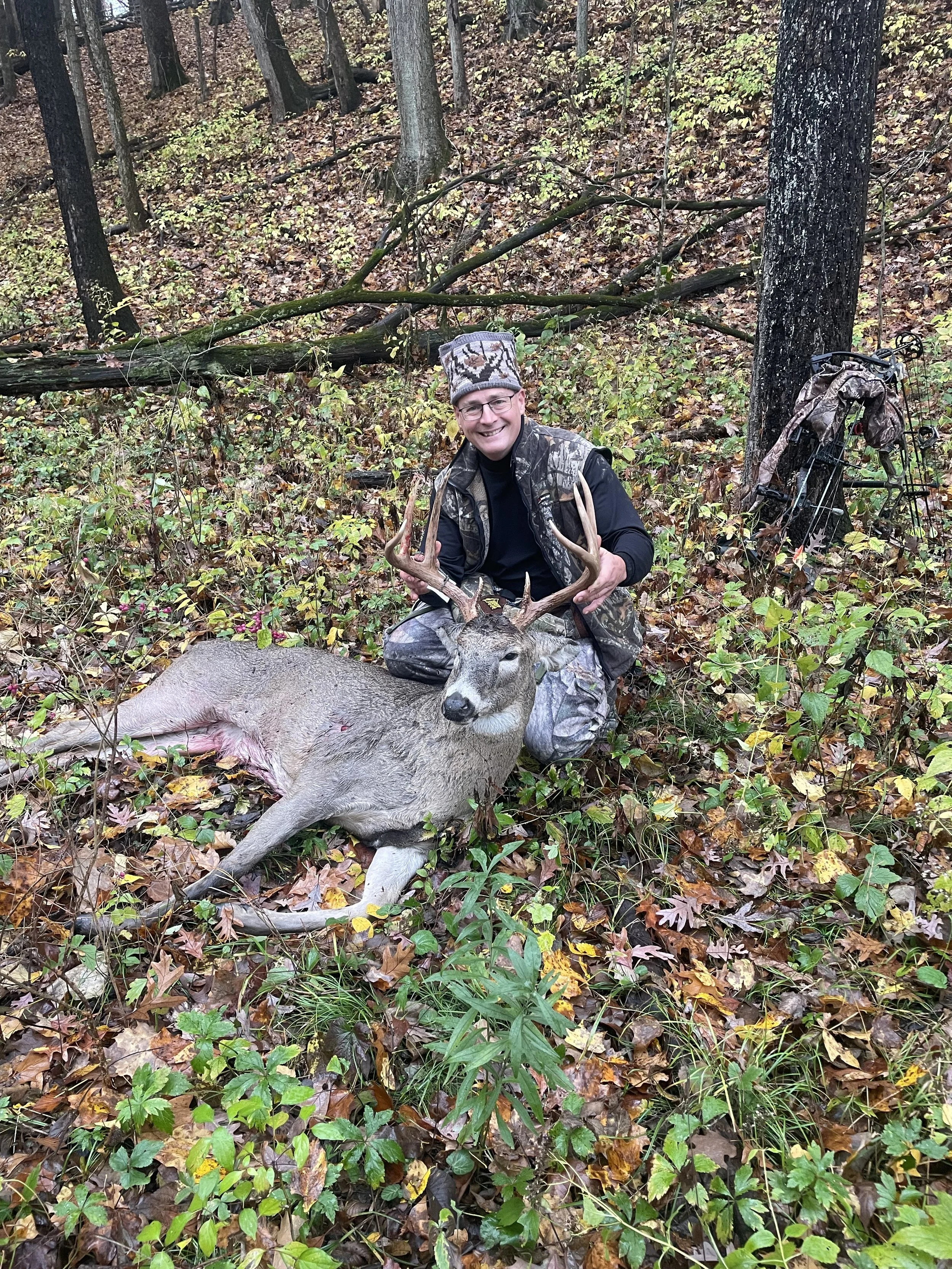 A man in camouflage and black clothing smiling and holding deer antlers while kneeling next to a dead deer in a wooded area with fallen leaves and trees.