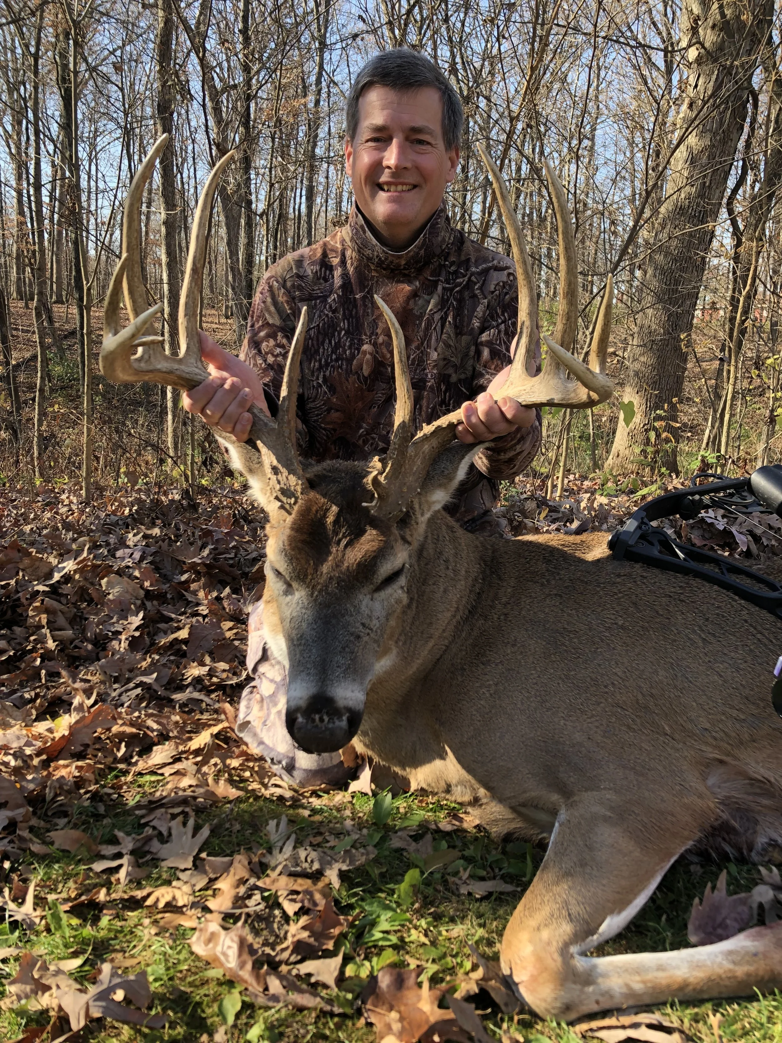 A man wearing camouflage clothes is smiling and holding the antlers of a large deer laying on the ground in a wooded area with leaf-covered ground and trees.