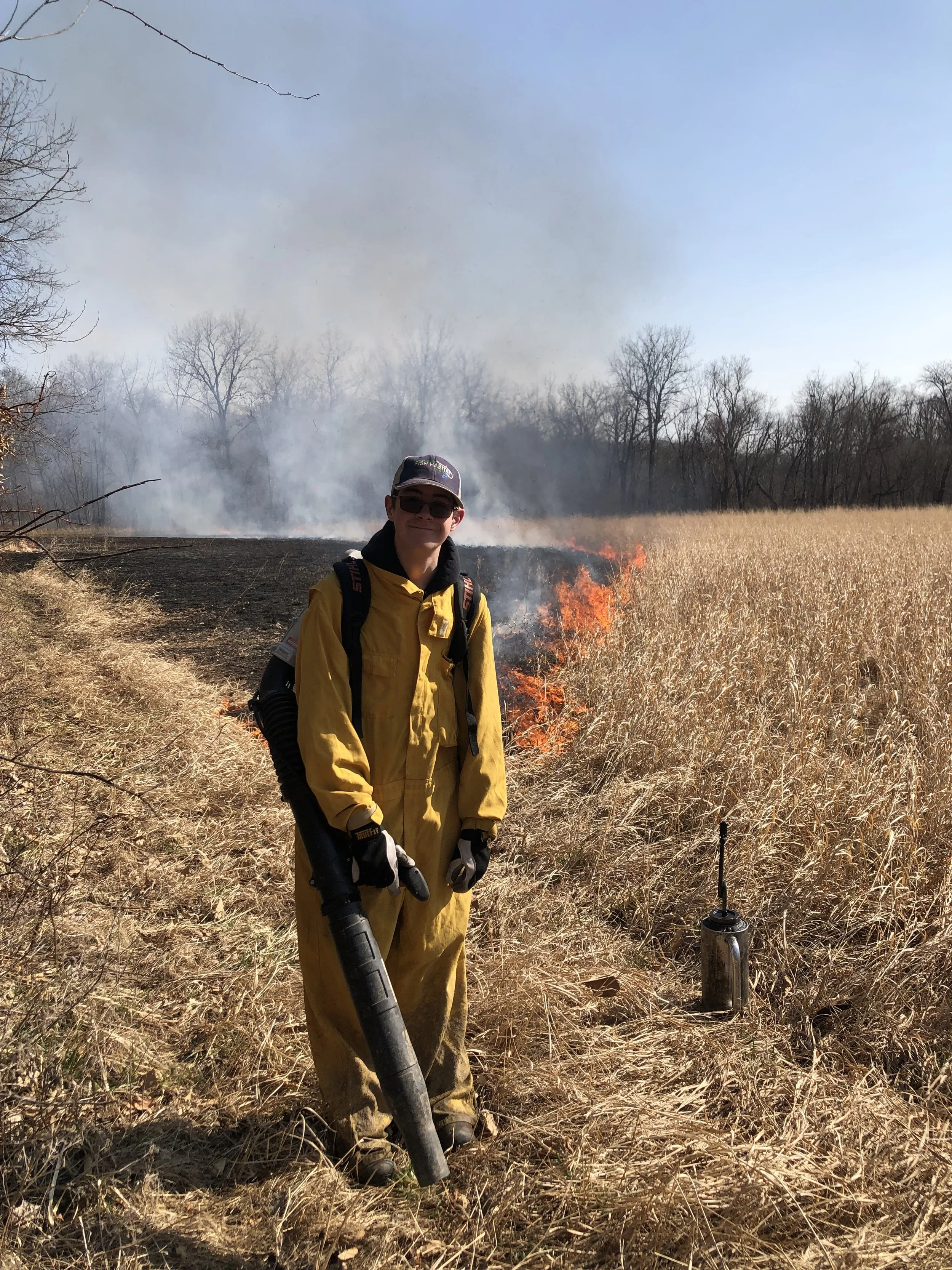 A firefighter stands in a field of dry grass and weeds, holding a leaf blower. Smoke from a recent fire can be seen in the background, with some flames still burning near the horizon. There are leafless trees in the distance under a clear blue sky.