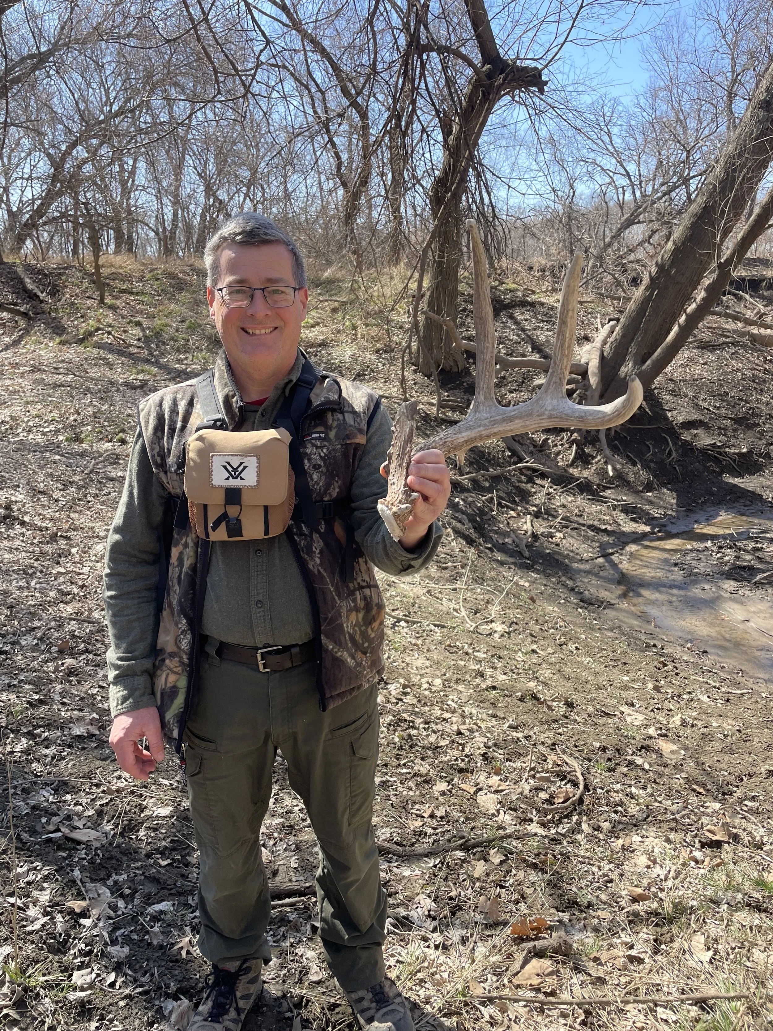 A man smiling and holding a shed deer antler in a wooded area with leafless trees and patches of sunlight.