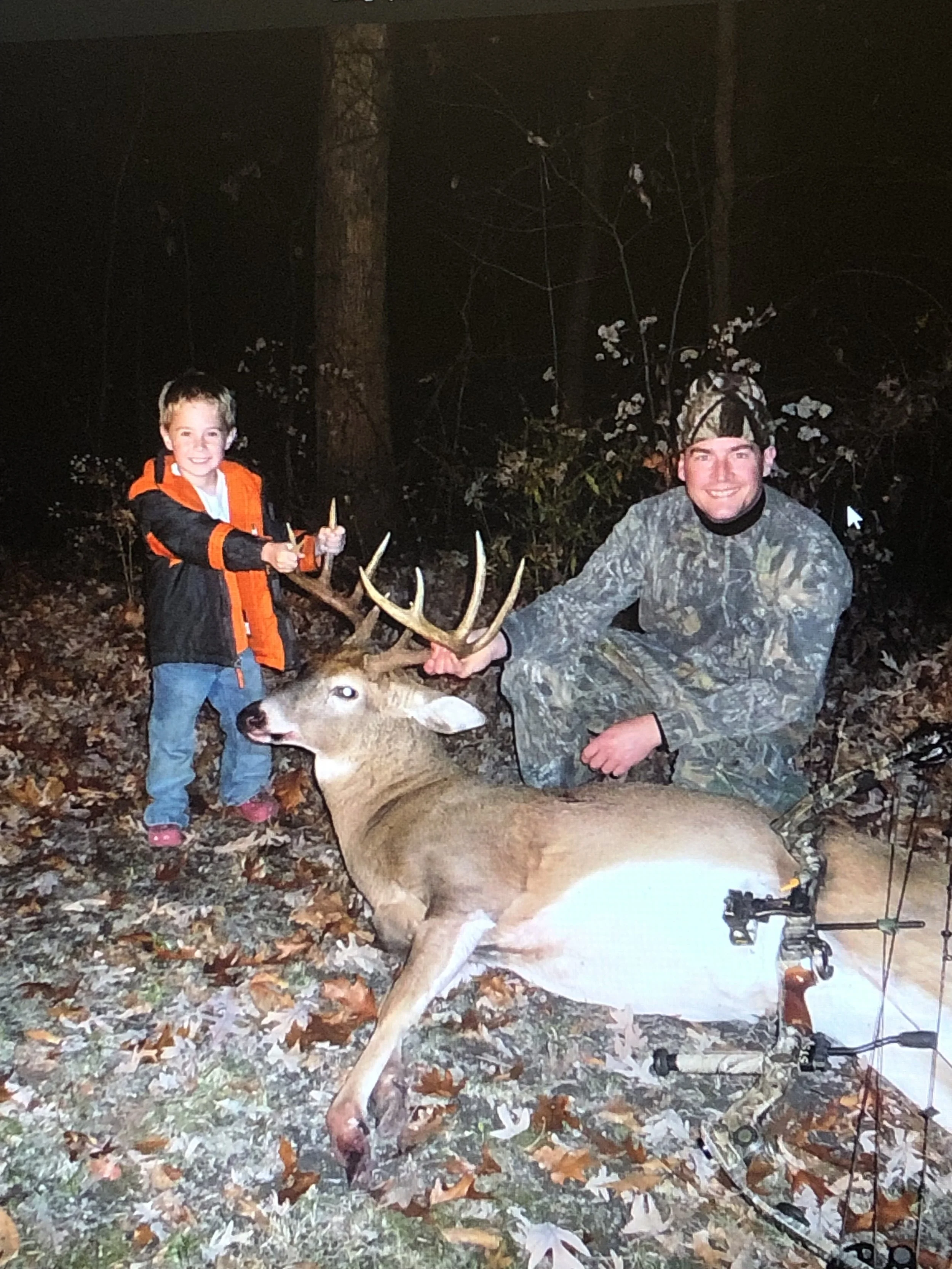 A young boy and a man outdoors at night in a wooded area, showing a recently caught deer with antlers. The boy is smiling and holding a knife, while the man, dressed in camouflage and wearing a headlamp, is kneeling beside the deer, which is lying on the ground.