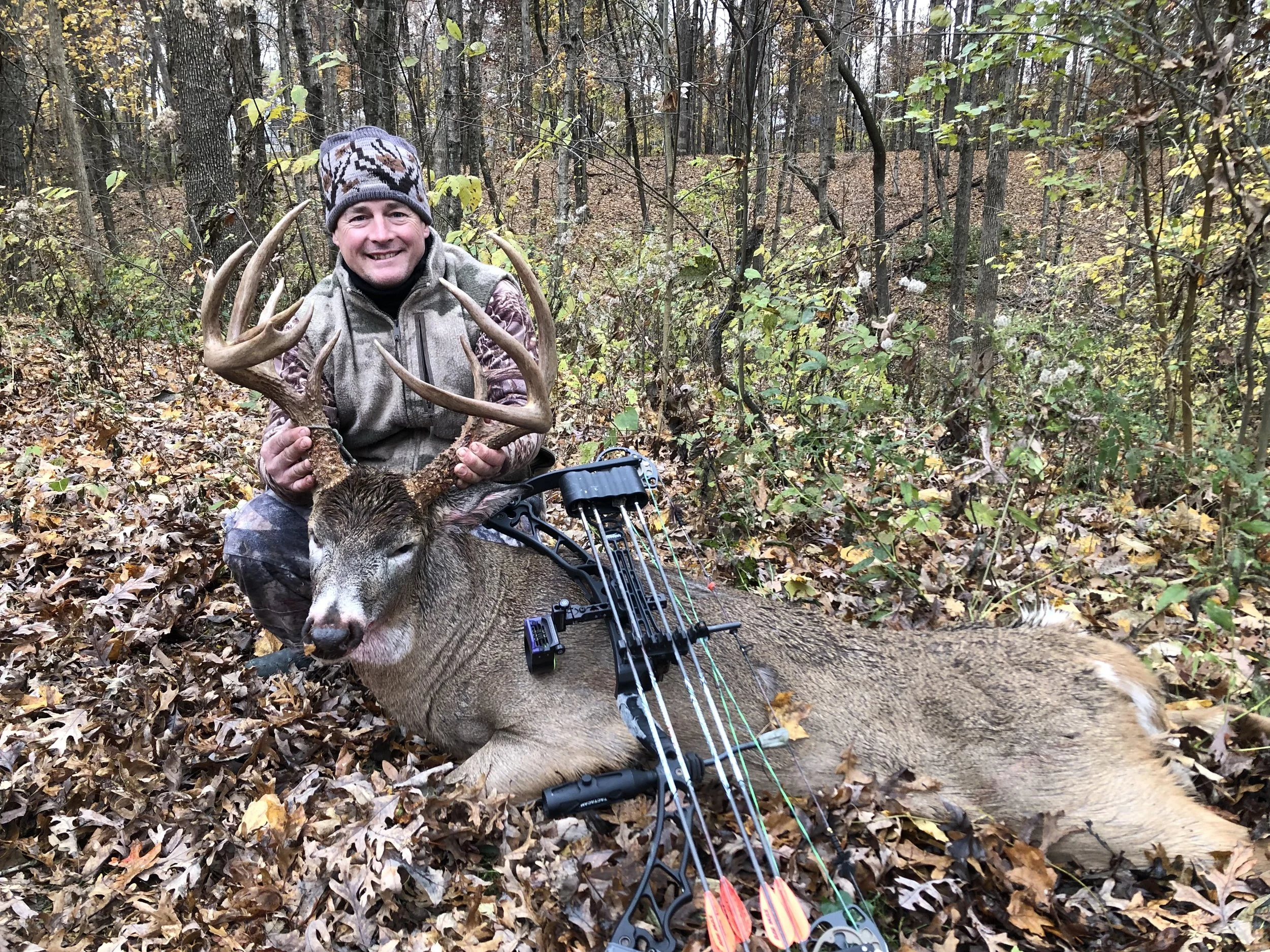 A man in camouflage clothing kneeling in a forest, holding the antlers of a deer he has hunted, with a bow and arrows lying in the leaves on the ground.