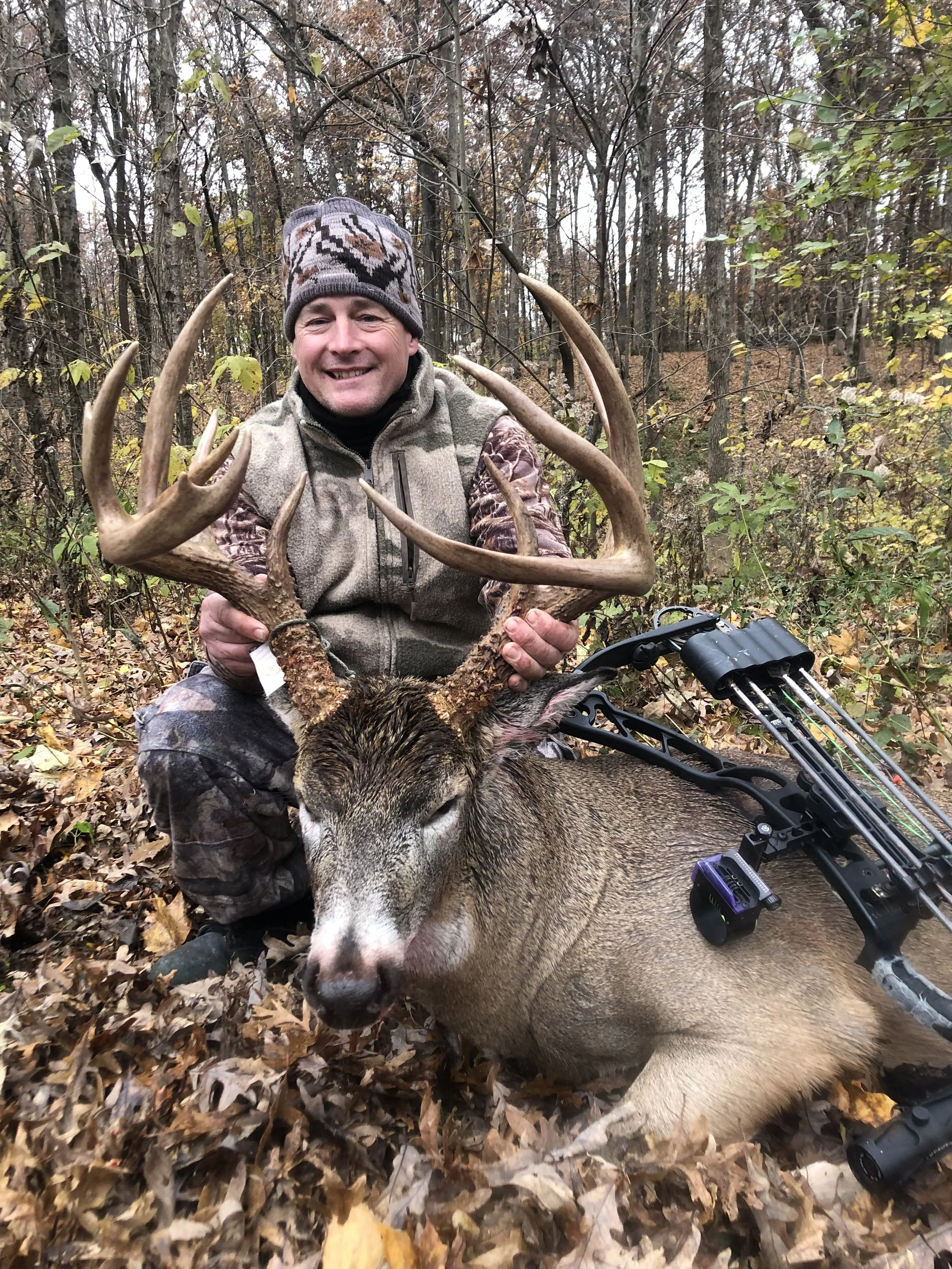 A man smiling and holding the antlers of a large deer he has hunted, sitting in a wooded area with fallen leaves, with a bow lying on the ground beside him.