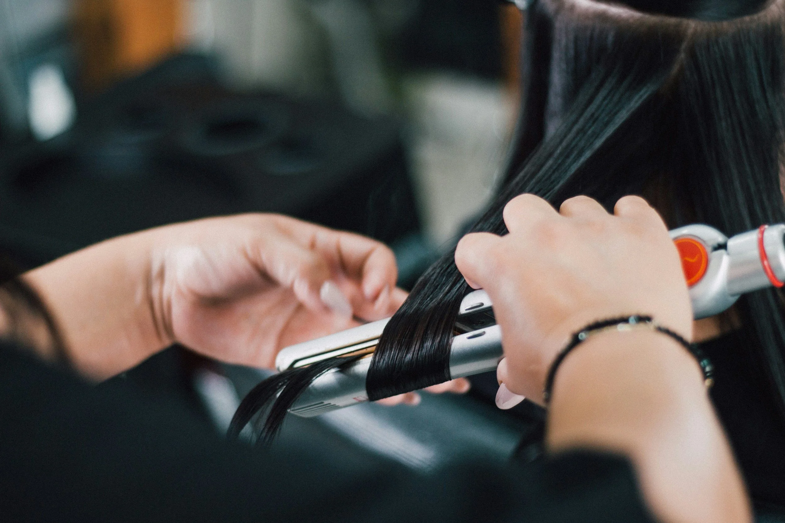 Close-up of a person curling or straightening their black hair with a flat iron, wearing a black bracelet.