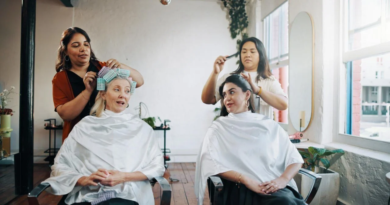 Two women getting hair and makeup done at a salon with stylists working on their hair.