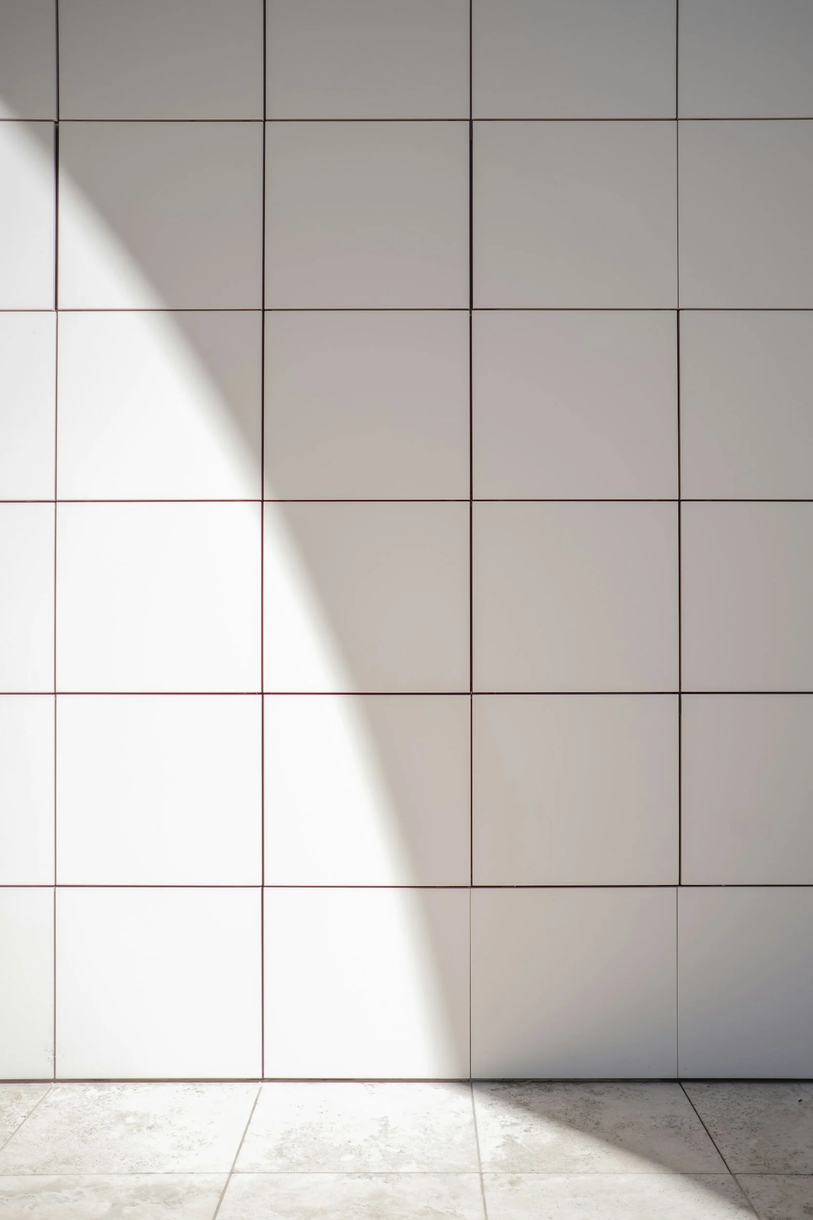 A tiled wall with square white tiles and visible grout lines, with sunlight casting a curved shadow on the left side of the wall, and a tiled floor with similar white tiles and light gray grout lines.